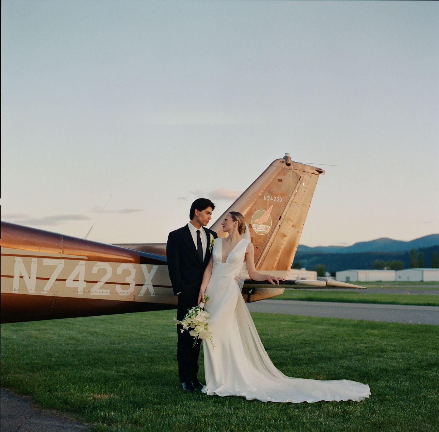 Bride and groom pose together in front of a vintage plane at sunset on their wedding day.