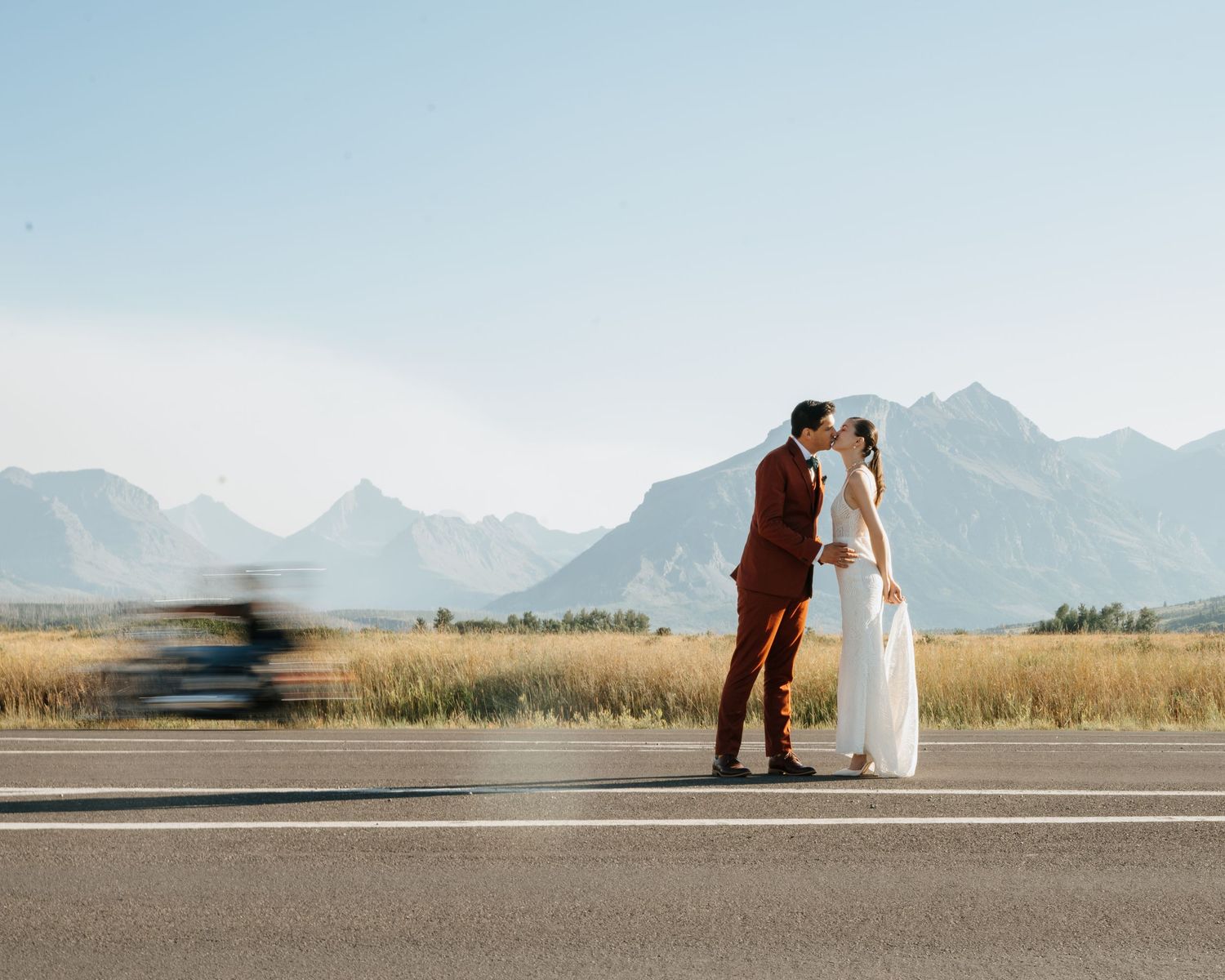 A bride and groom embrace on a road with mountains in the background.