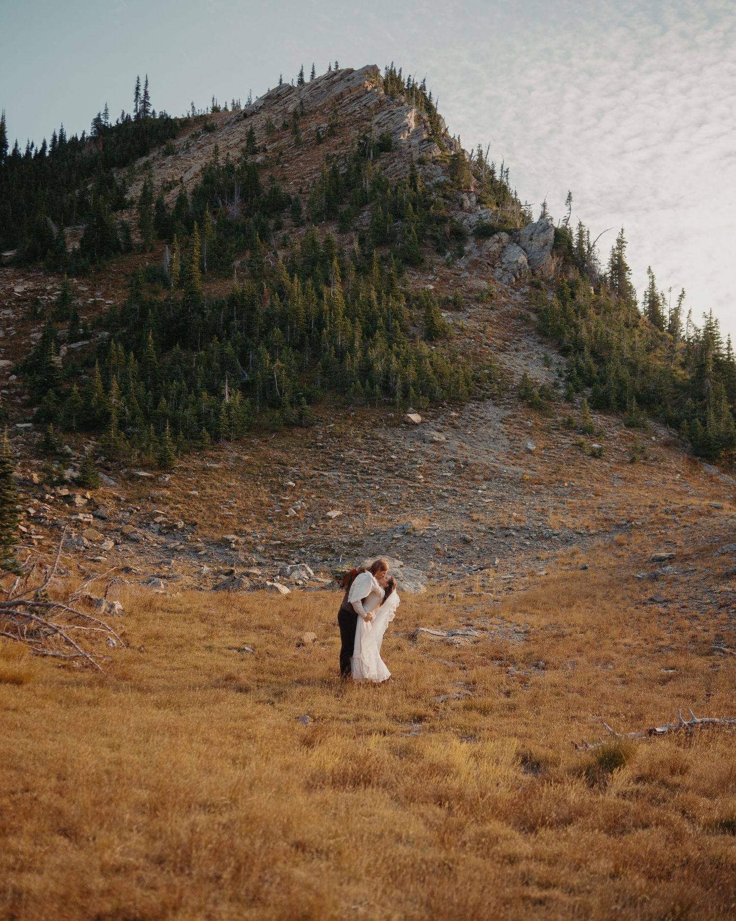 A couple shares an intimate moment on a rocky mountain slope surrounded by evergreen trees.