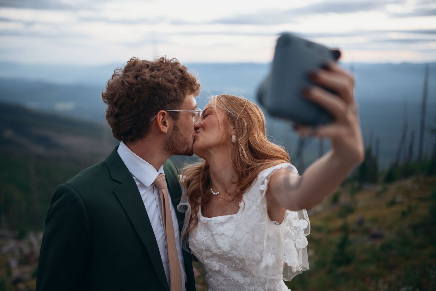 A couple shares a romantic kiss while taking a selfie against a scenic mountain landscape at sunset.