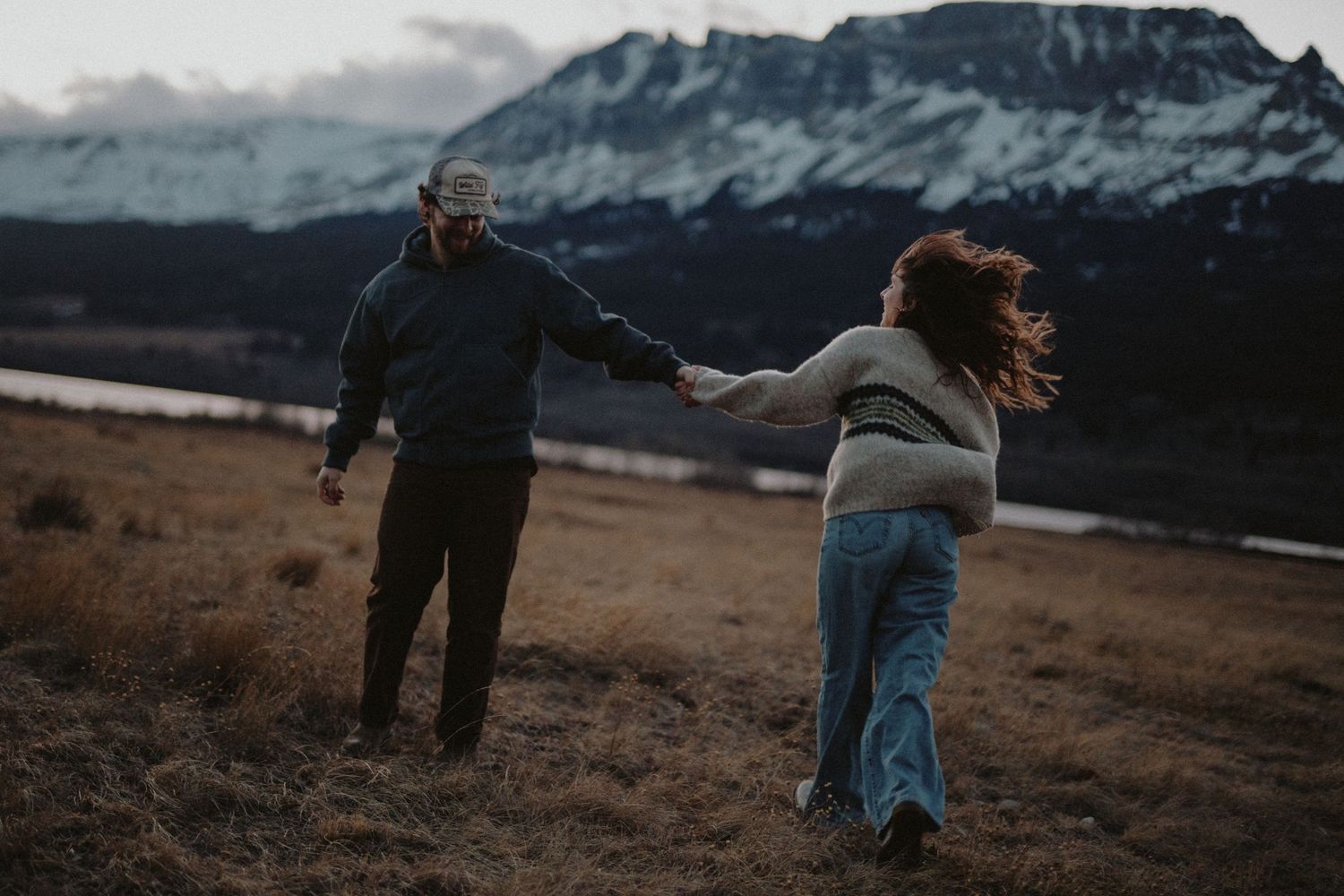 A couple holding hands while walking in a scenic, mountainous landscape on an overcast day.