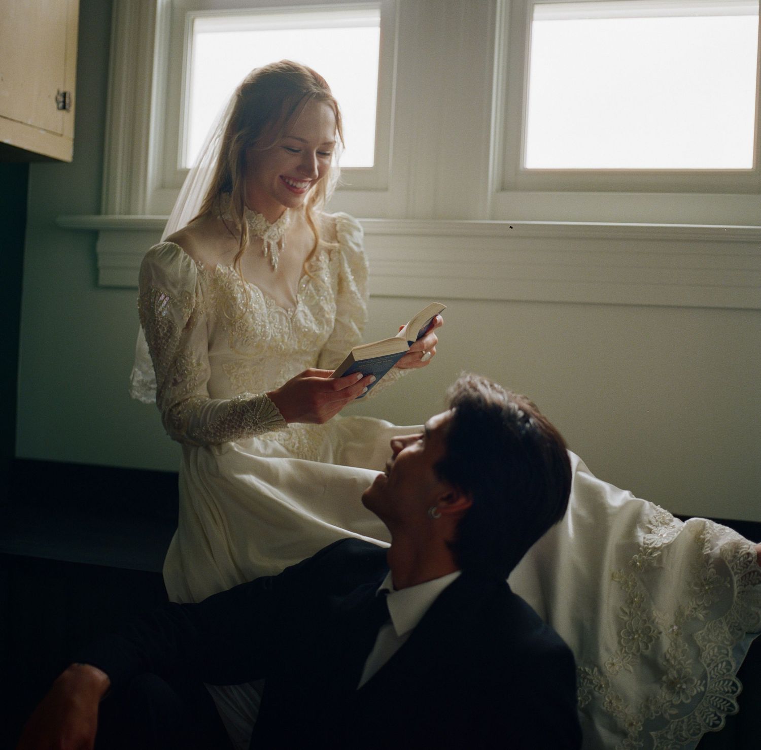 Bride in lace wedding dress reads to groom in suit seated on floor near bright windows.