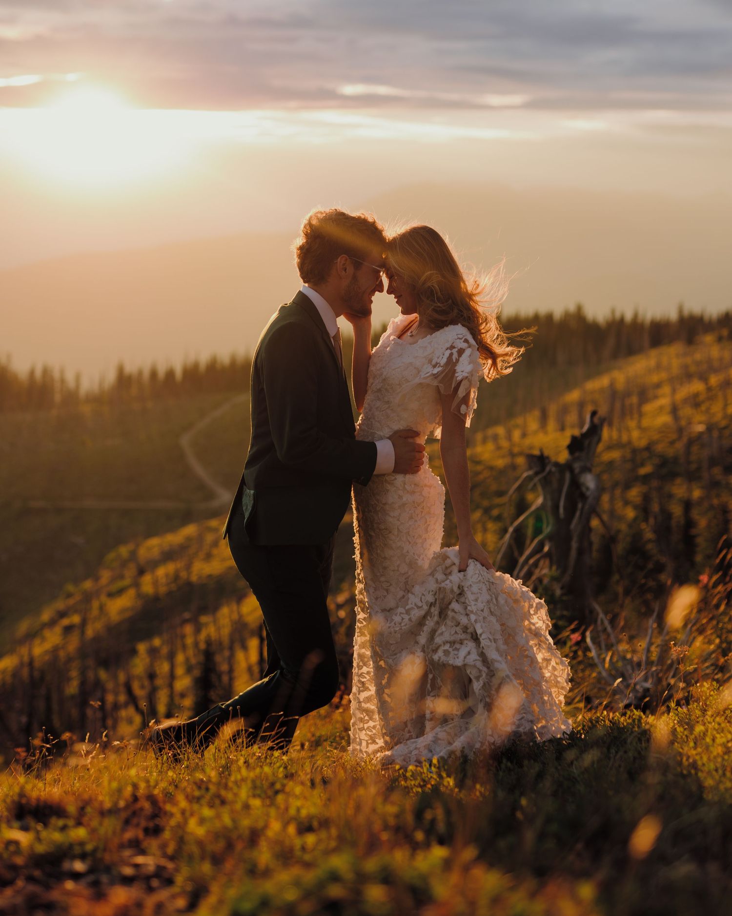 A couple in wedding attire shares an intimate moment at sunset on a scenic mountain hillside.