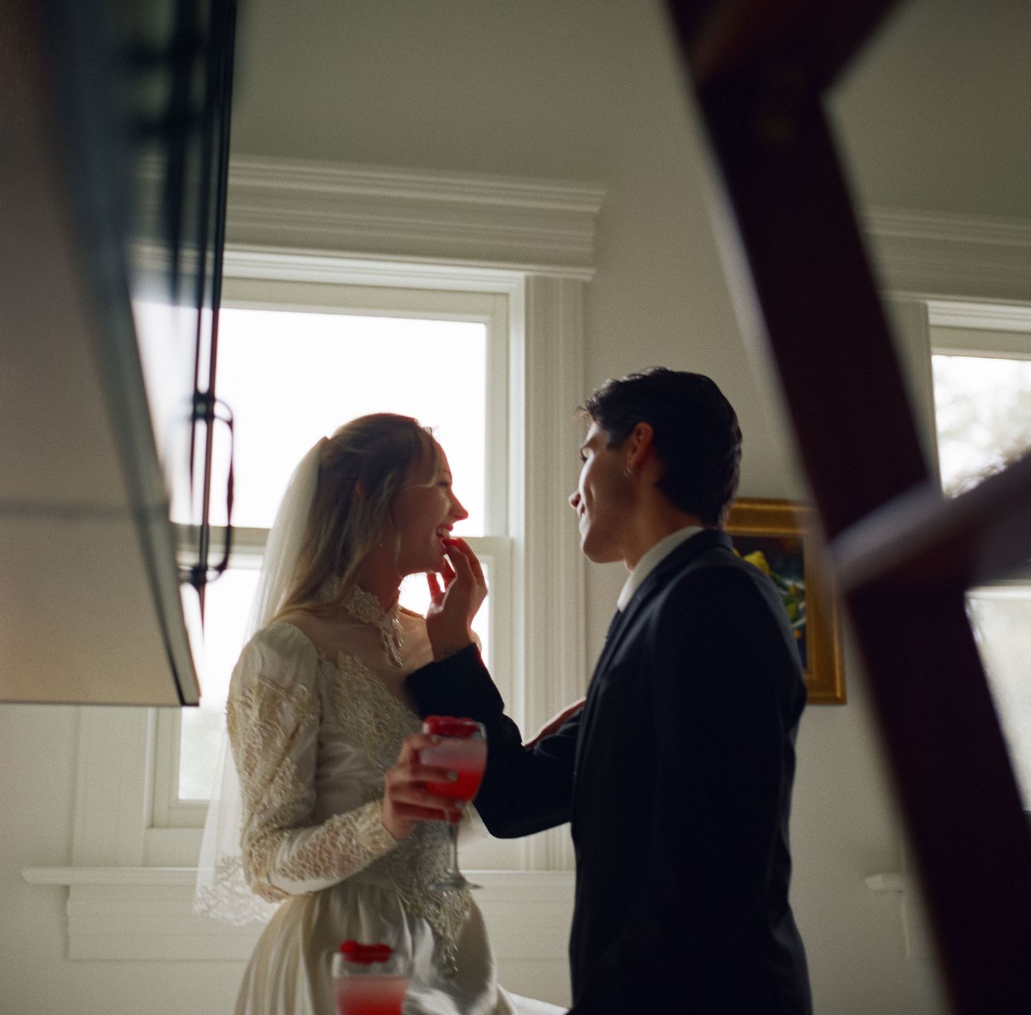 A bride in lace gown and groom in dark suit share an intimate moment by a window at their wedding reception.