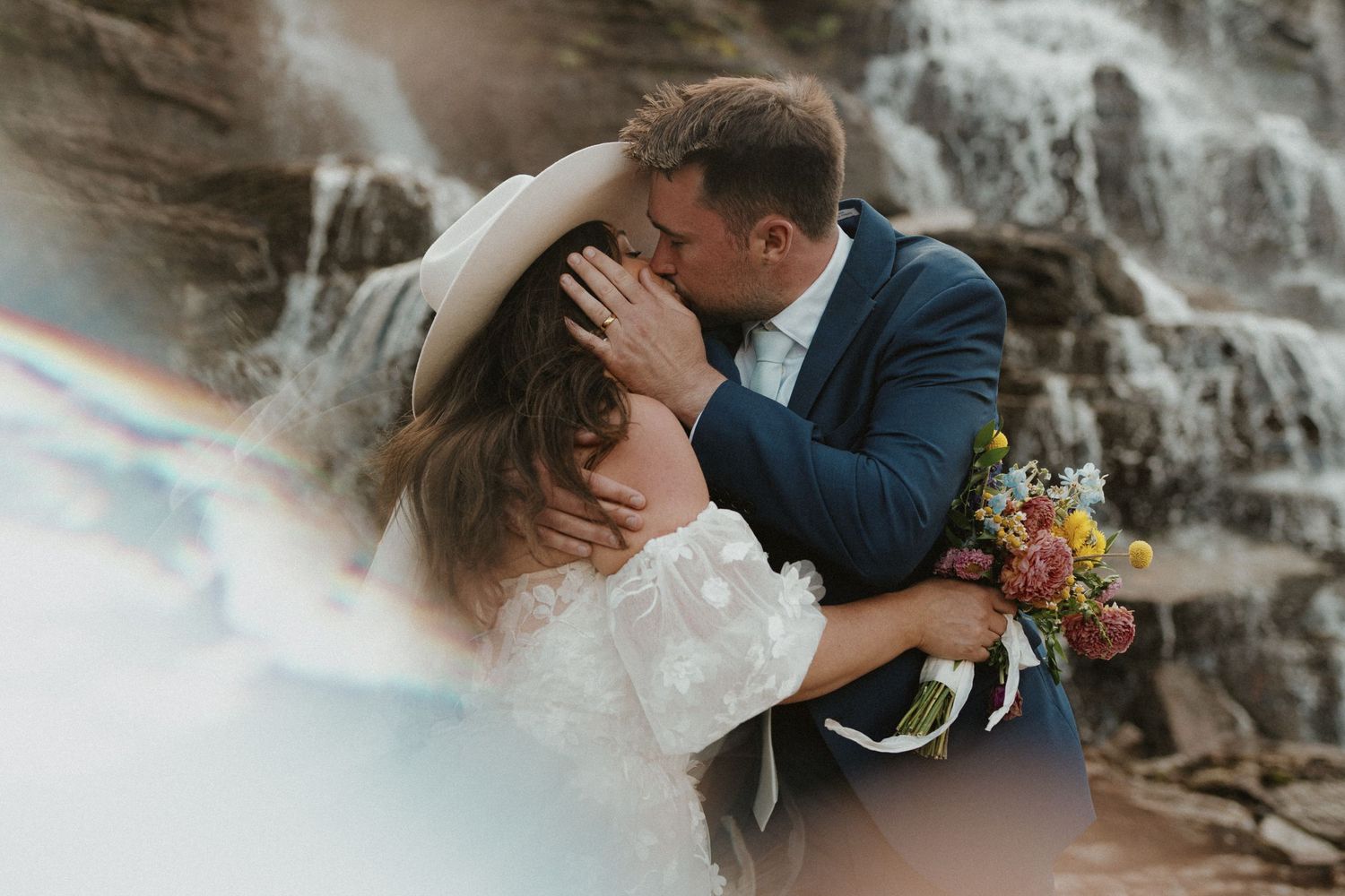 A romantic wedding couple shares a kiss in front of a cascading waterfall with a bouquet of autumn flowers.