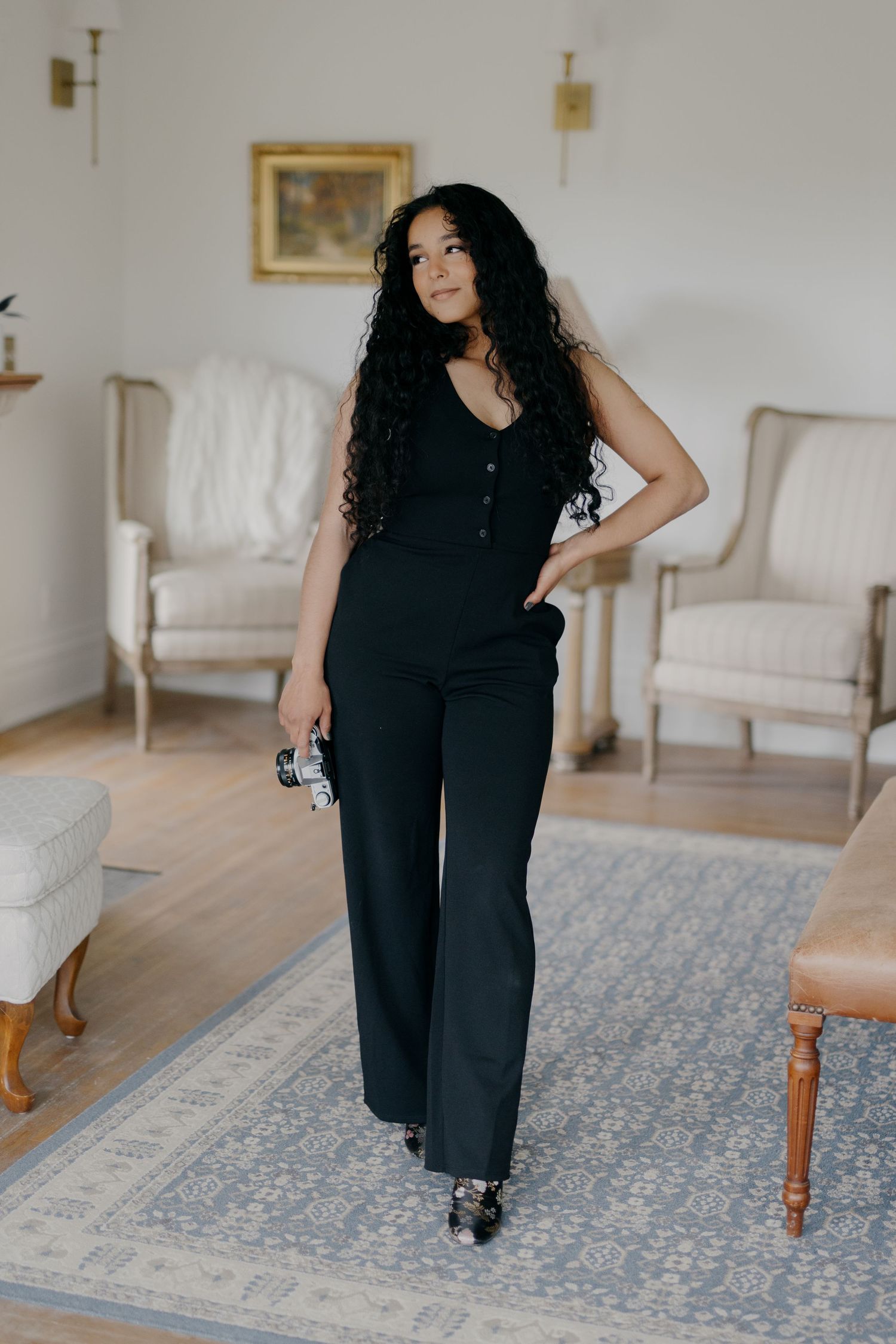 A person in a black outfit poses in a minimalist room with white walls, vintage furniture and a blue patterned rug.