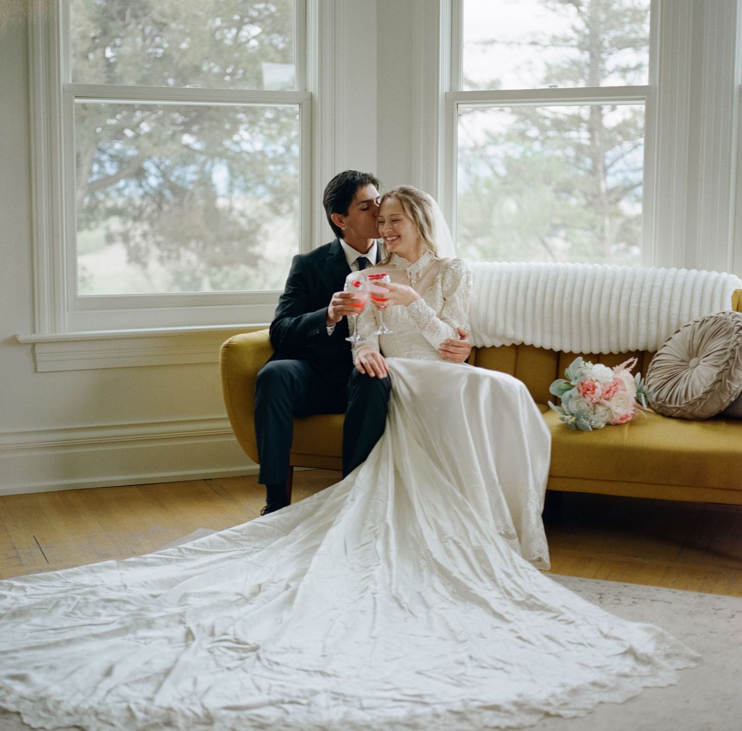 Bride and groom sitting on a yellow sofa, sharing a tender kiss near bright windows on their wedding day.