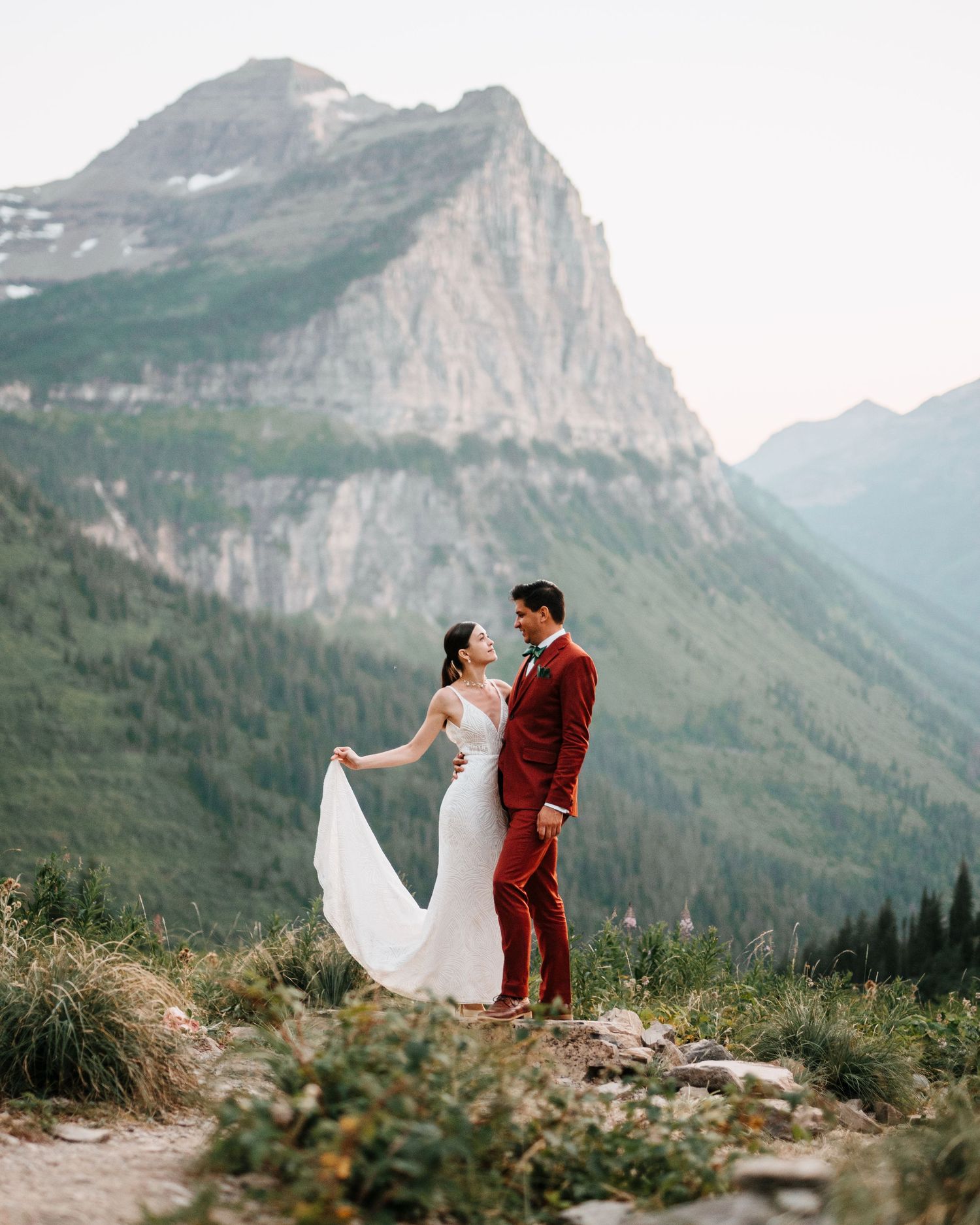 Couple in wedding attire stands on mountain summit with dramatic peaks and valleys of Glacier National Park in background.