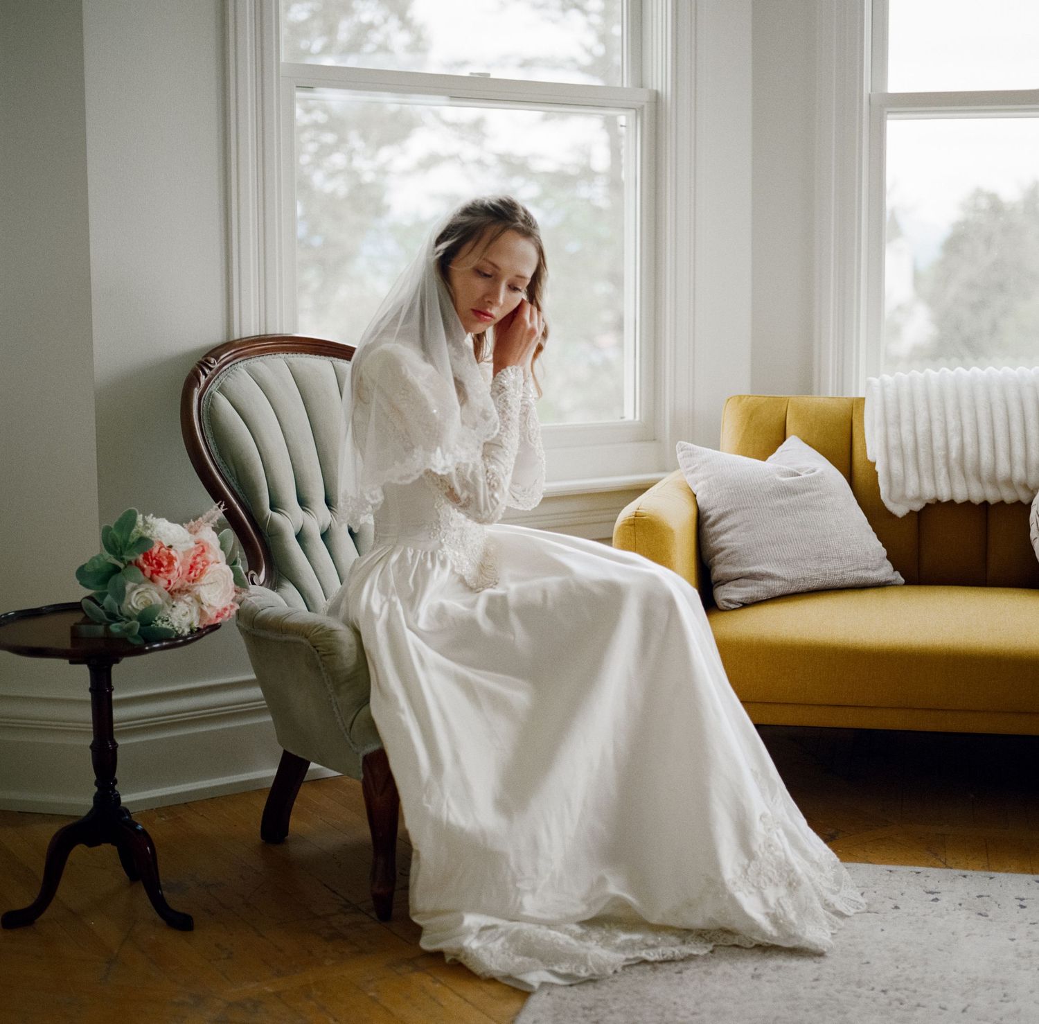A woman in a white wedding dress sitting in a chair by a window in a cozy room.