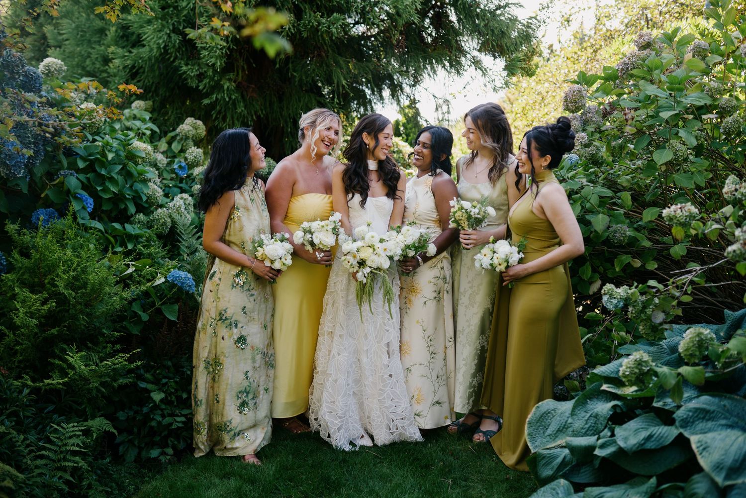 Bride in white gown with bridesmaids in yellow dresses holding white bouquets in a lush garden setting.