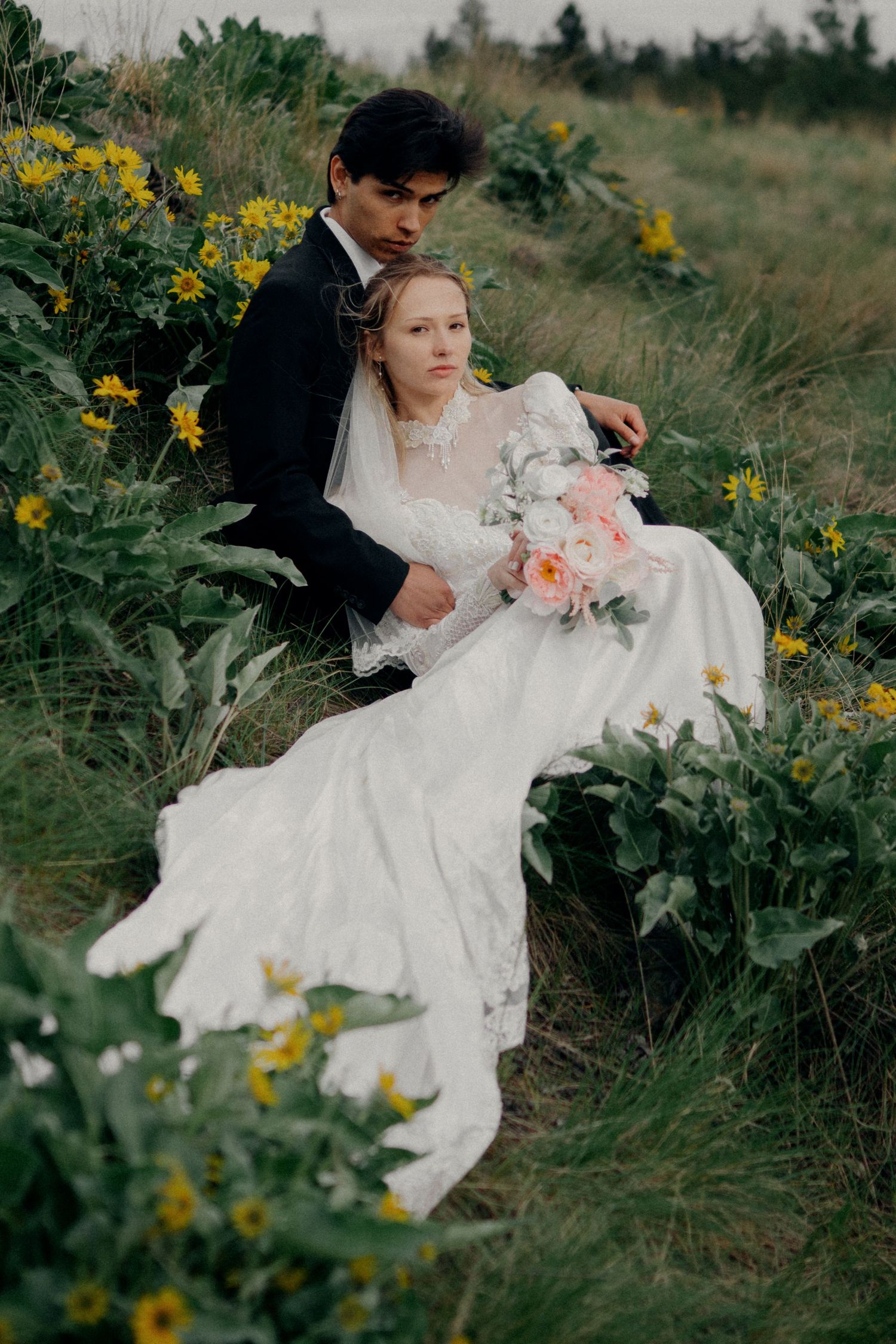 A couple in wedding attire reclines in a meadow filled with yellow wildflowers and green foliage.