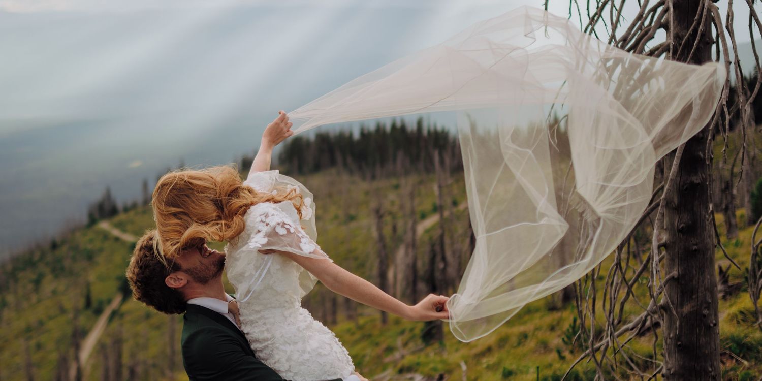 A bride's veil flows dramatically in the wind against a misty mountain landscape backdrop.