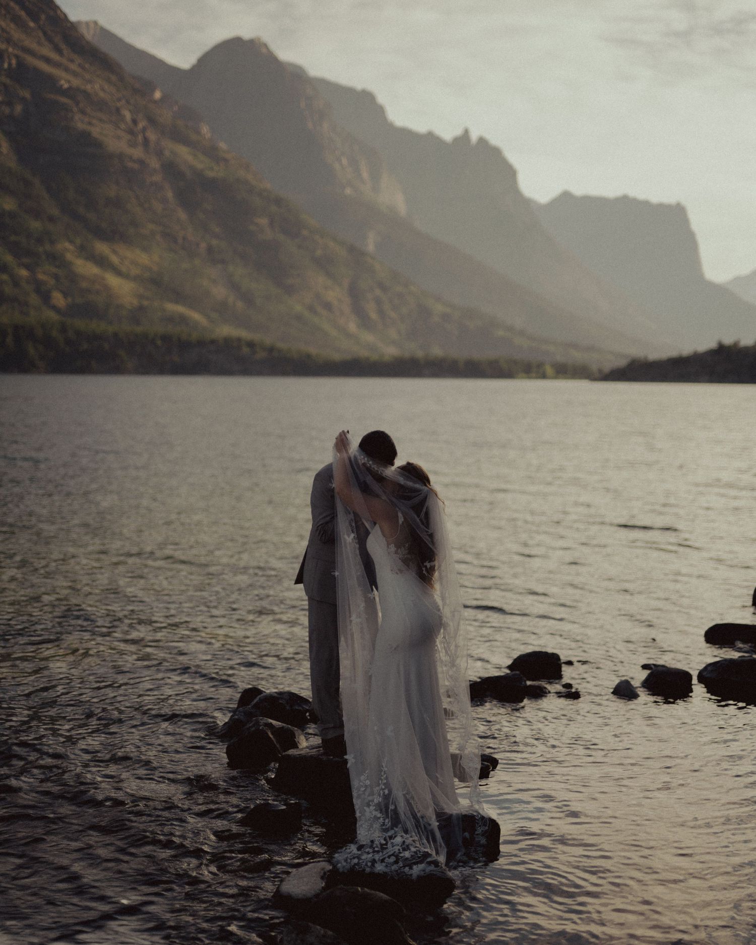 A couple in wedding attire stands at the edge of a misty lake surrounded by dramatic mountain scenery at sunset.