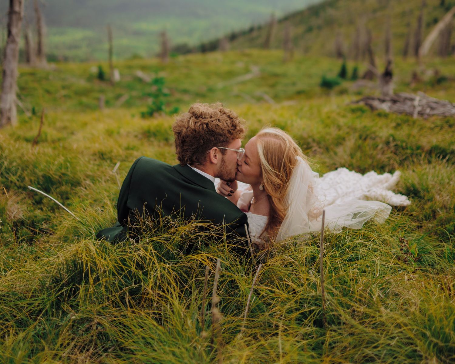 Couple lying in tall green grass during a romantic outdoor photoshoot in a scenic mountain landscape.