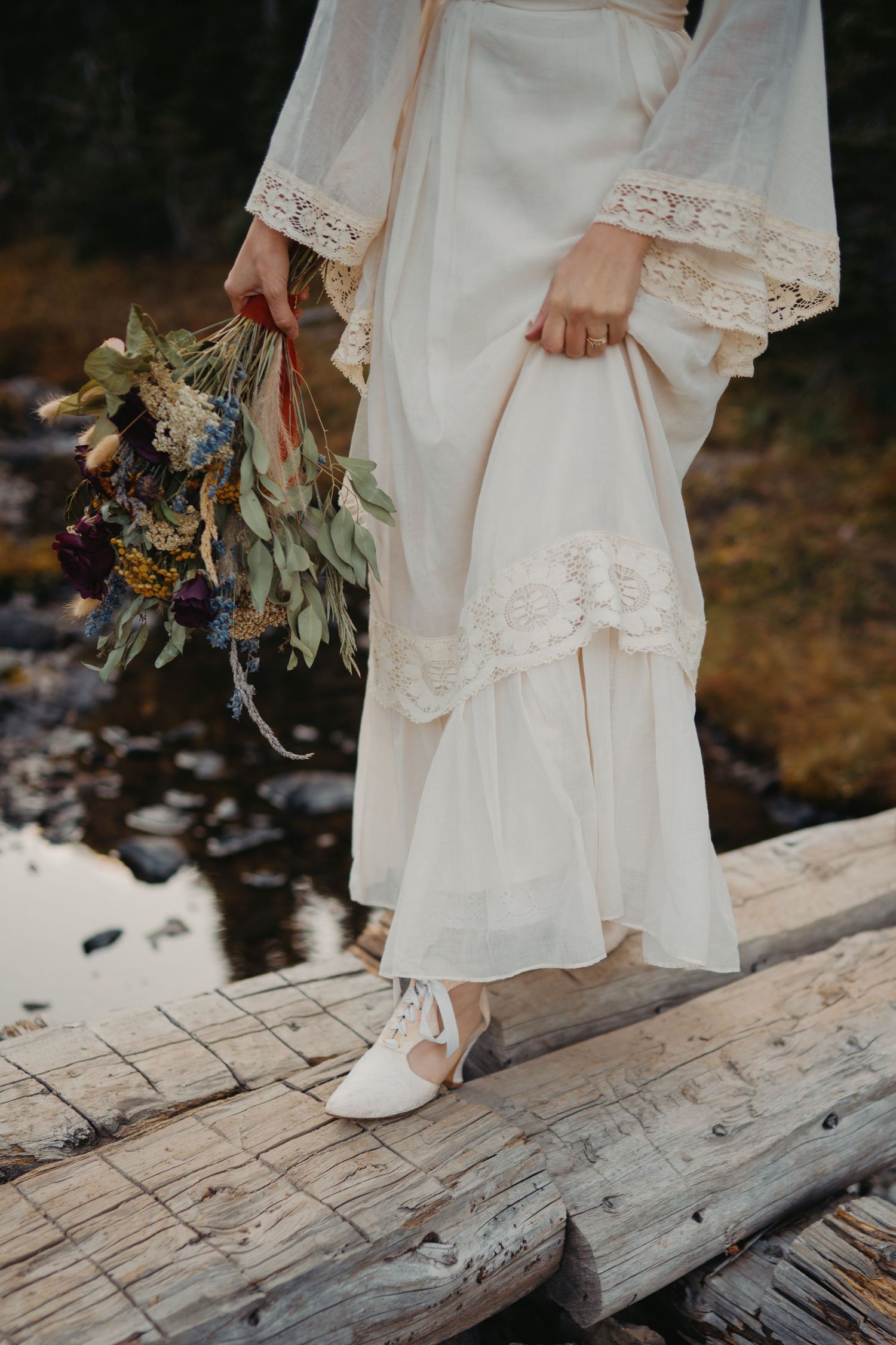 Close-up view of bride's white lace dress and bouquet while standing on rustic wooden planks in nature.