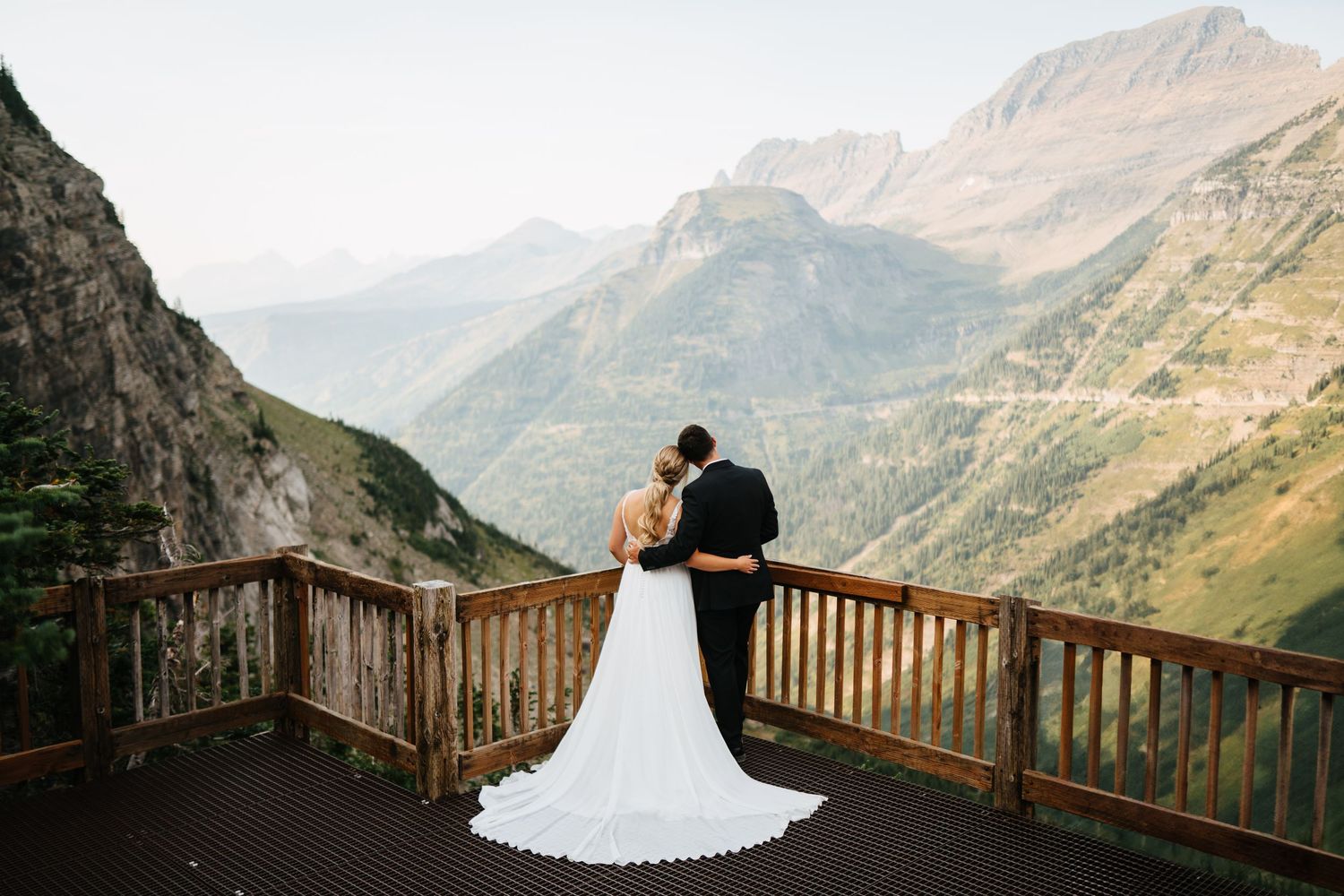 A couple in wedding attire embraces on a rustic wooden deck overlooking majestic mountain peaks at sunset.