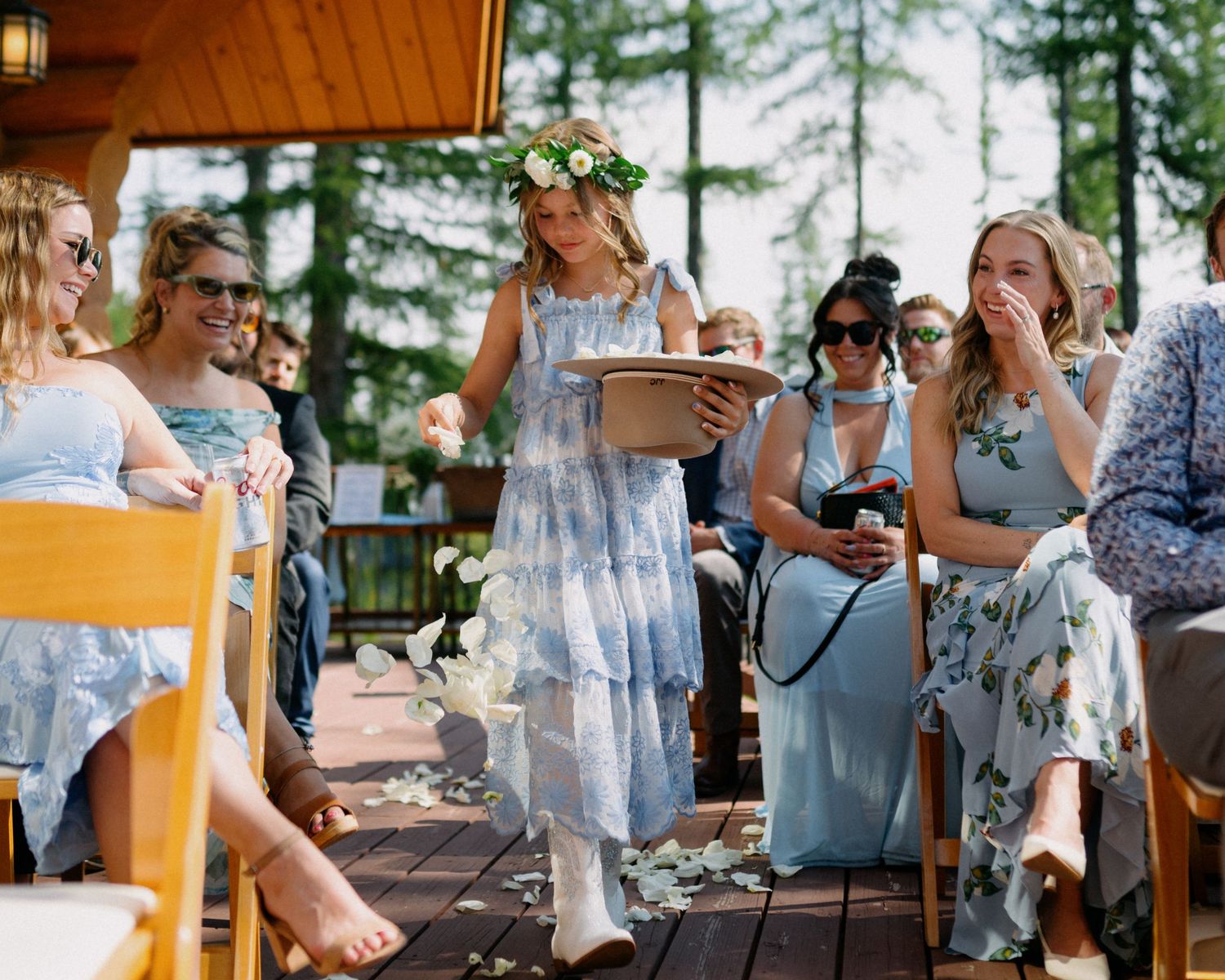 Flower girl in floral dress and flower crown walks down outdoor wedding aisle as guests smile and cry.