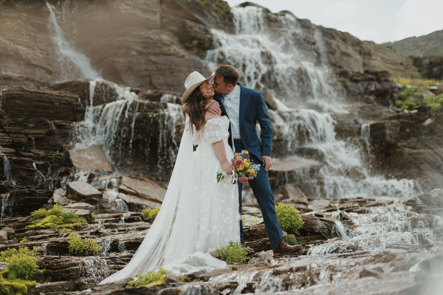 A couple in wedding attire poses near a cascading waterfall on their special day.