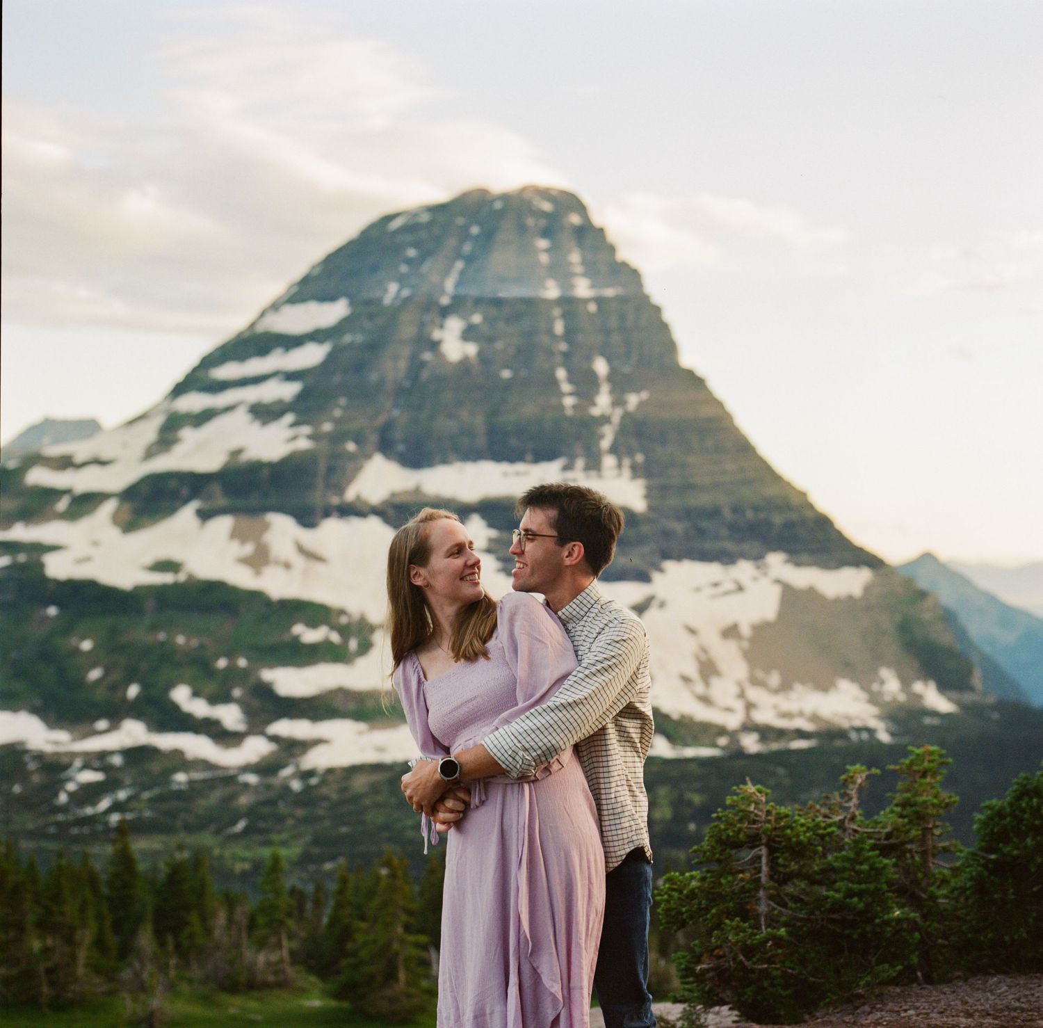 Couple embracing with snowy mountain peak backdrop during golden hour at Glacier National Park engagement session.