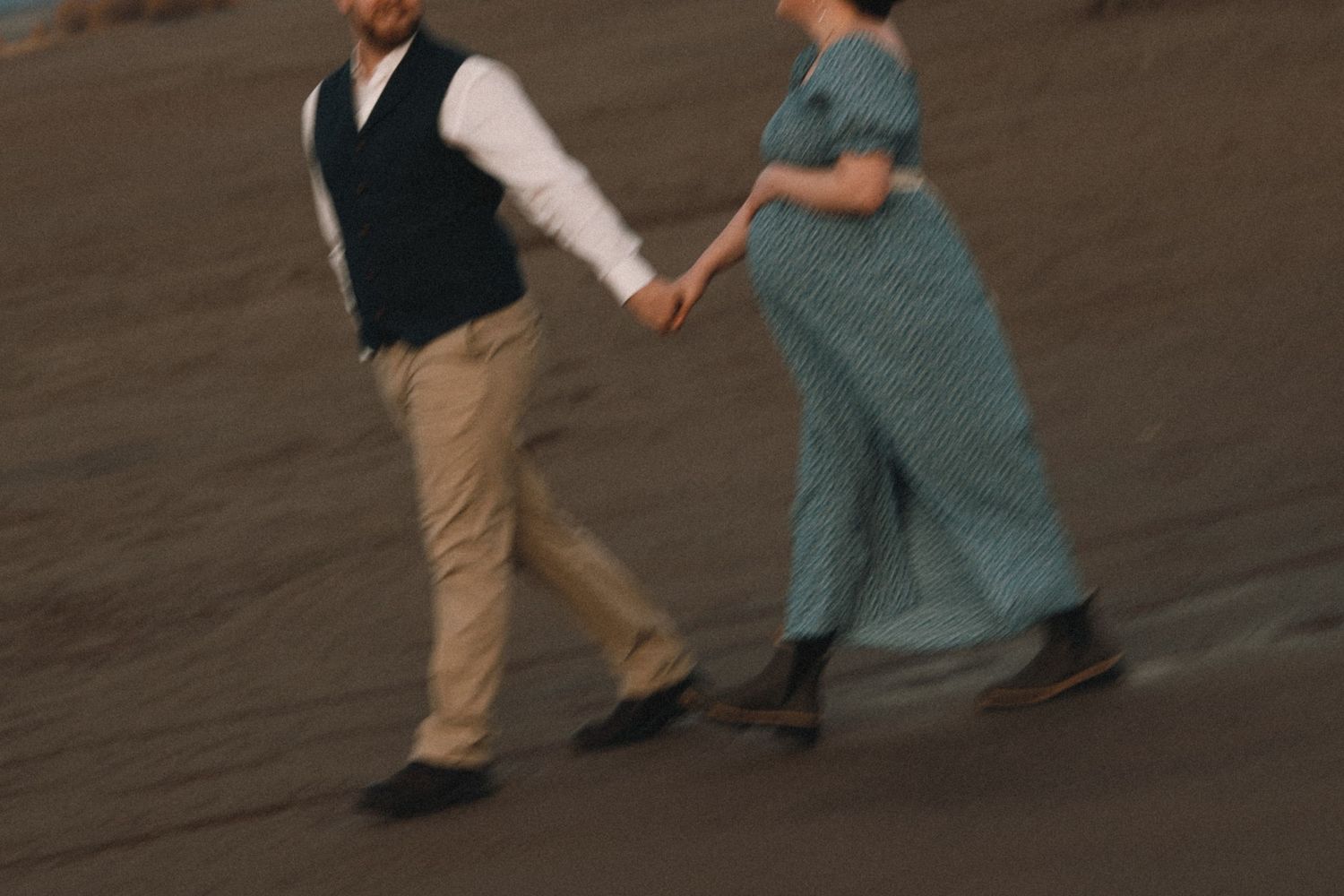 A pregnant woman in a teal dress holds hands with a man in a vest as they walk together on a sandy beach.