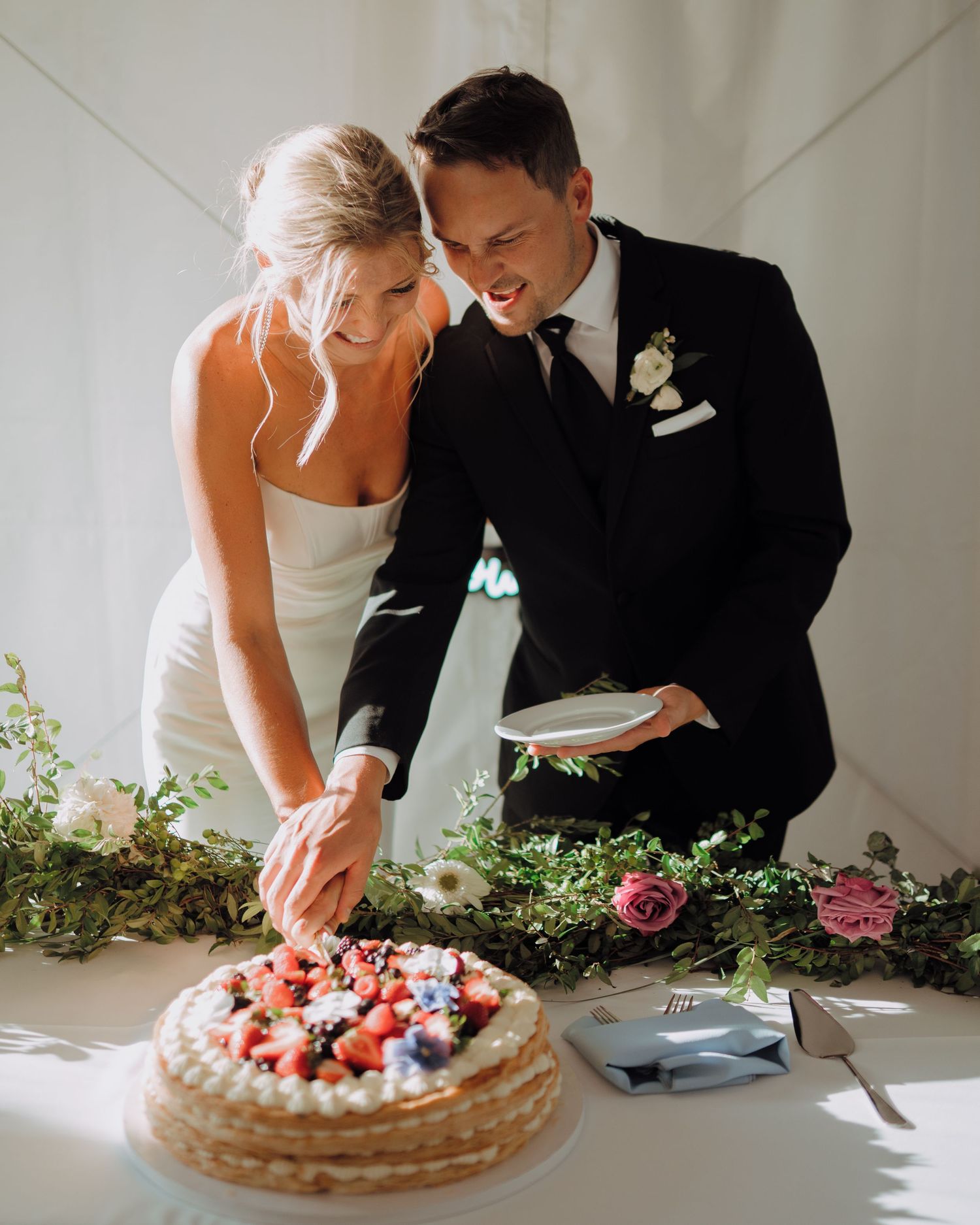 A couple in wedding attire leans over a table to cut their berry-topped cake in a bright, minimalist setting.