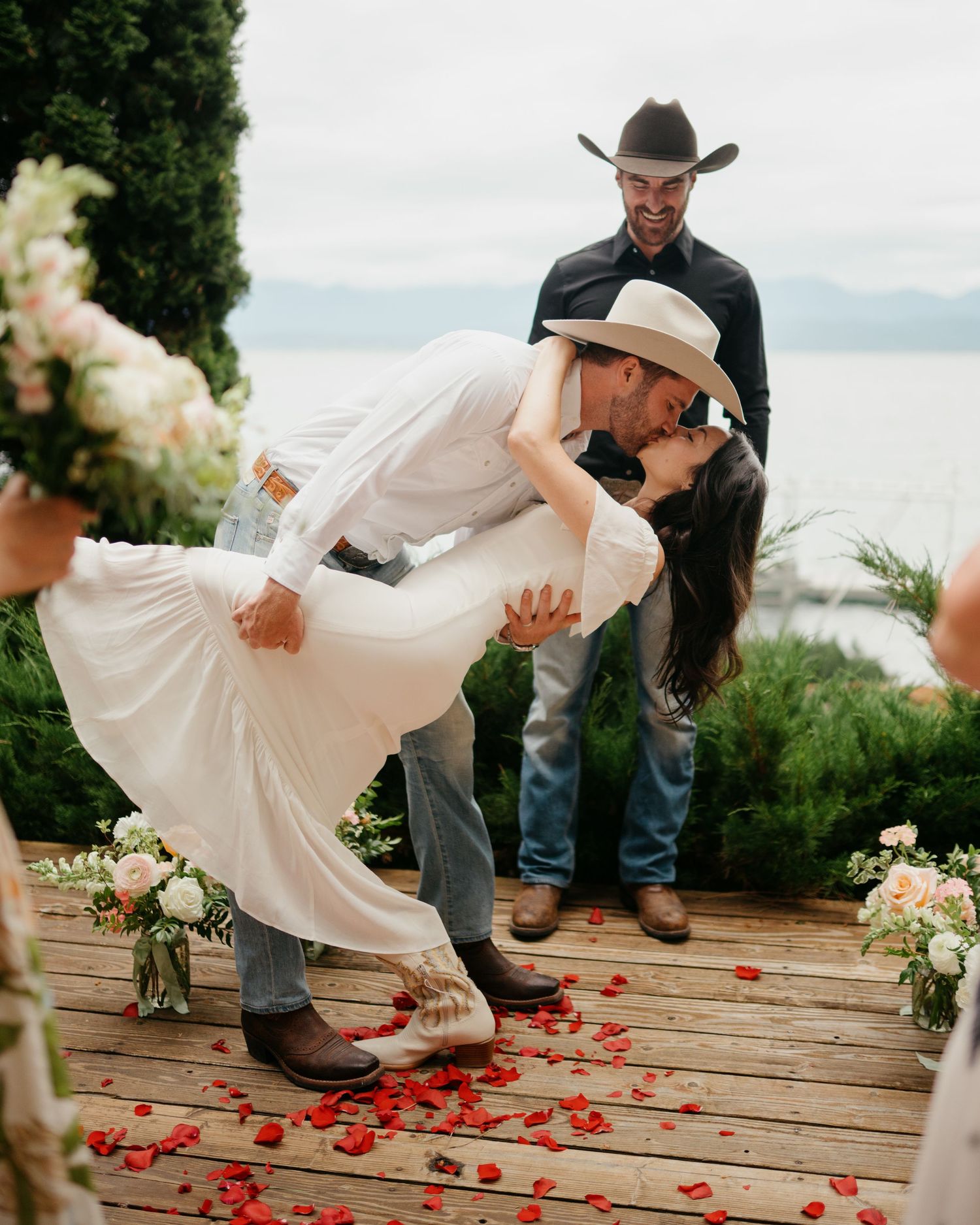 A romantic wedding kiss on a wooden deck decorated with red rose petals and white floral arrangements by the water.