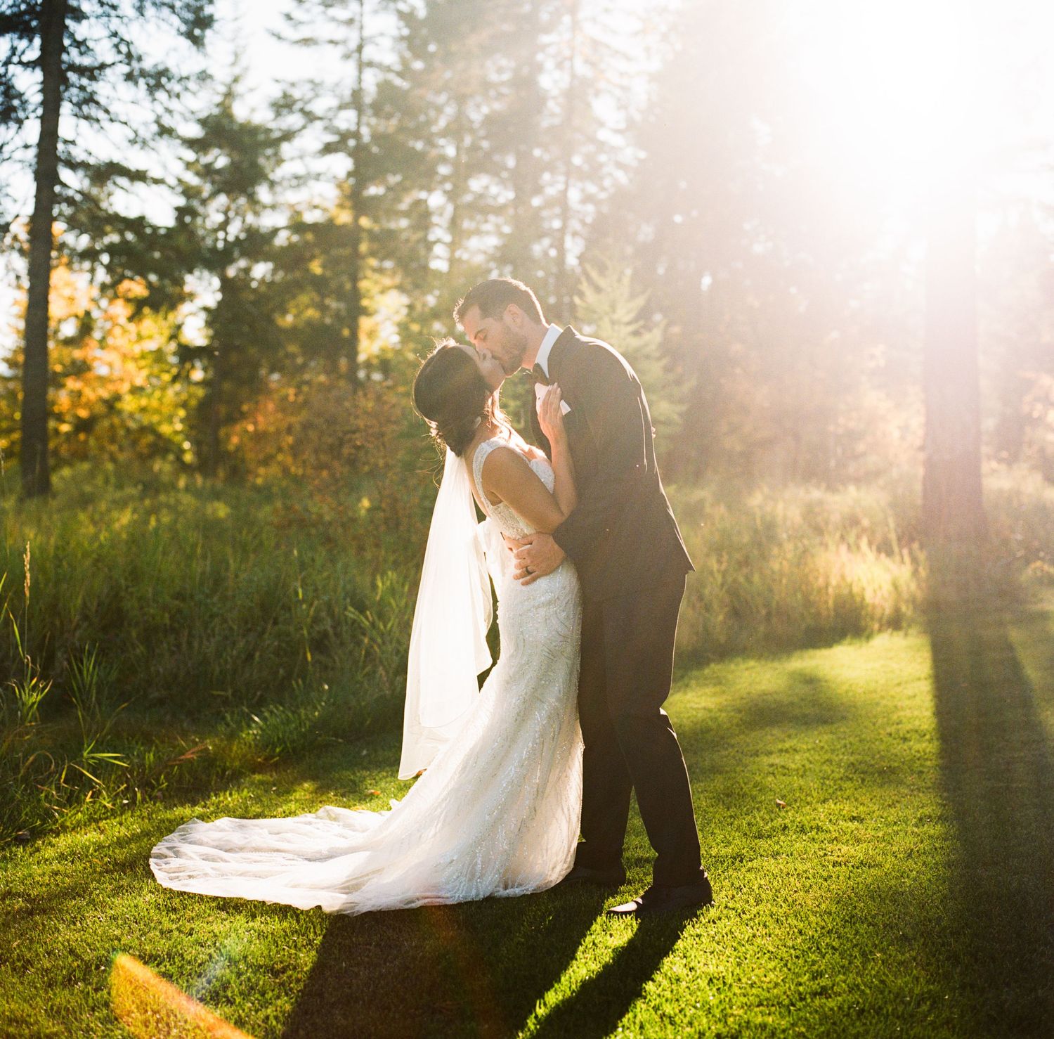 A bride and groom share a romantic kiss in a sunlit forest clearing, with golden autumn trees glowing behind them.