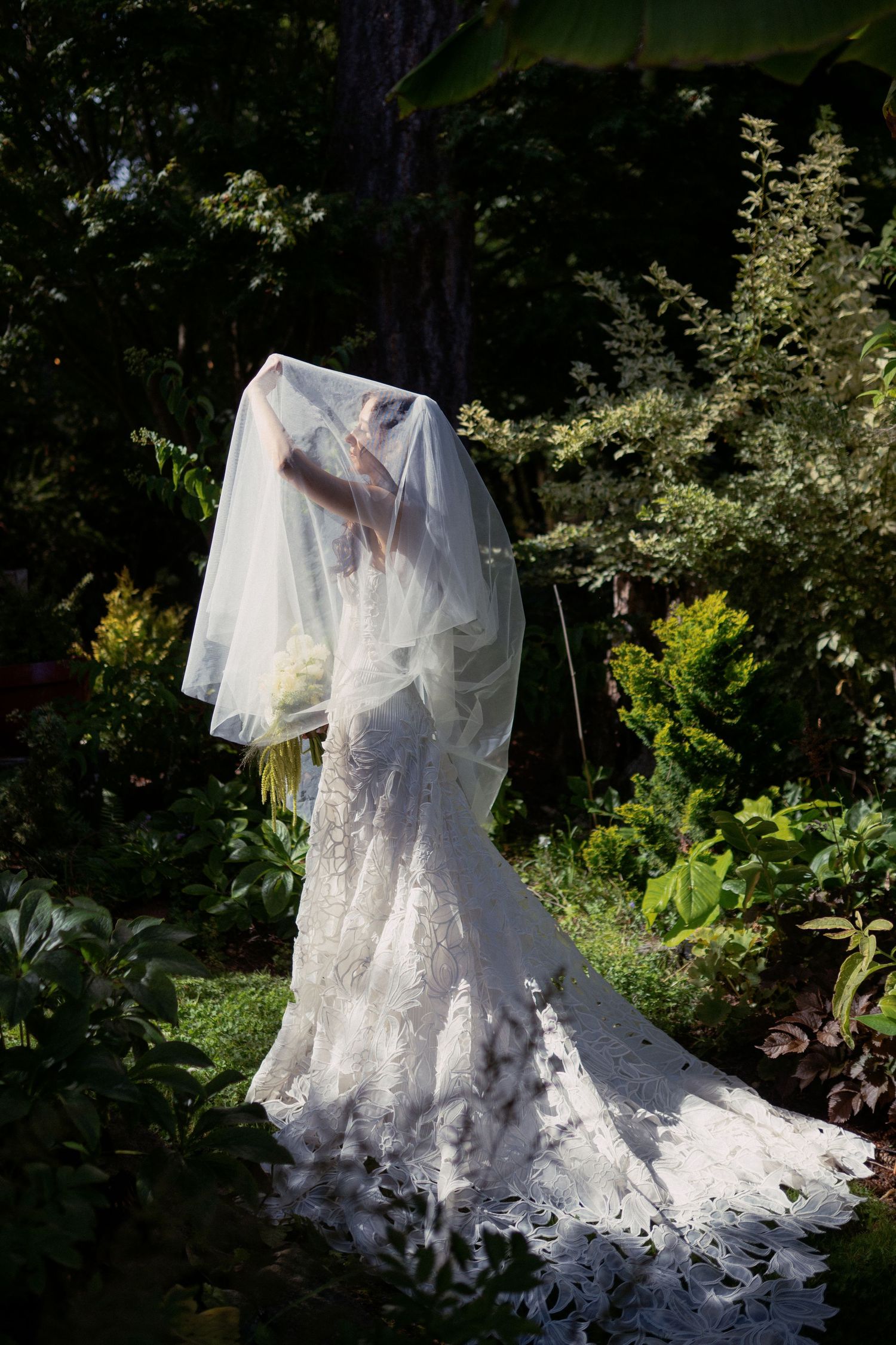 Bride in lace wedding gown and veil standing in lush garden, holding bouquet, with sunlight filtering through trees.