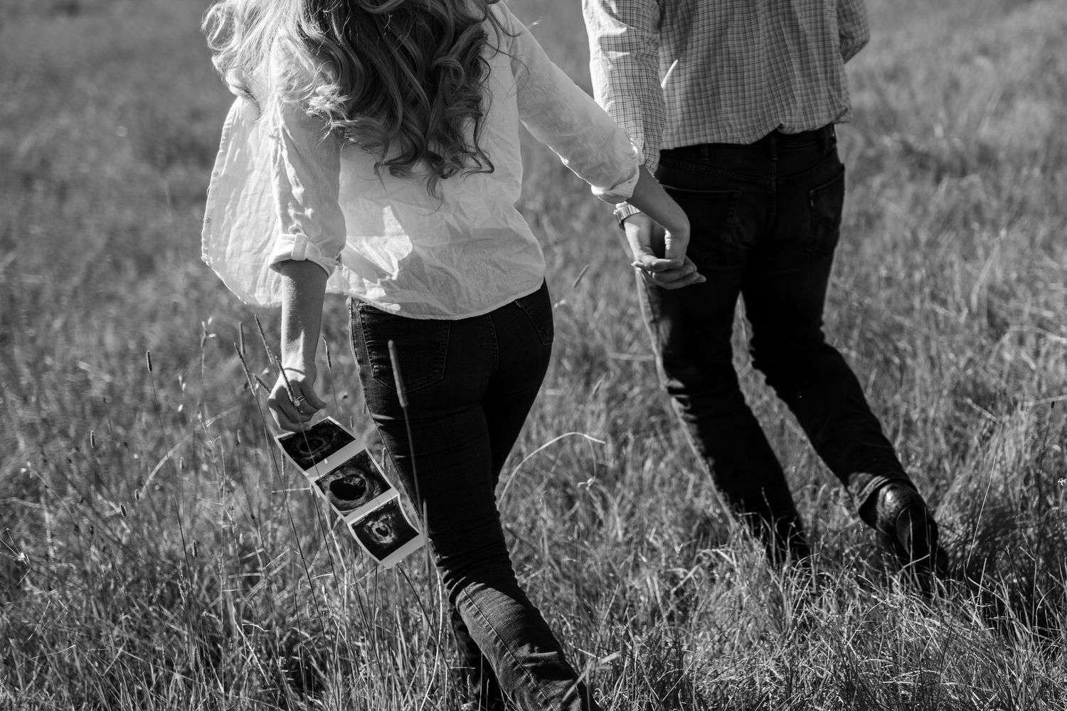 Black and white photo showing people's legs and feet walking through tall grass in a field.