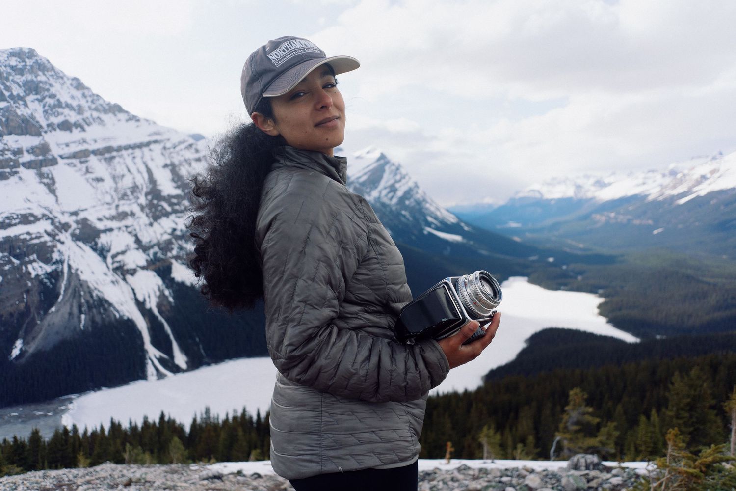 Woman holding camera at mountain overlook with snow-capped peaks, evergreen forest, and turquoise lake in background.
