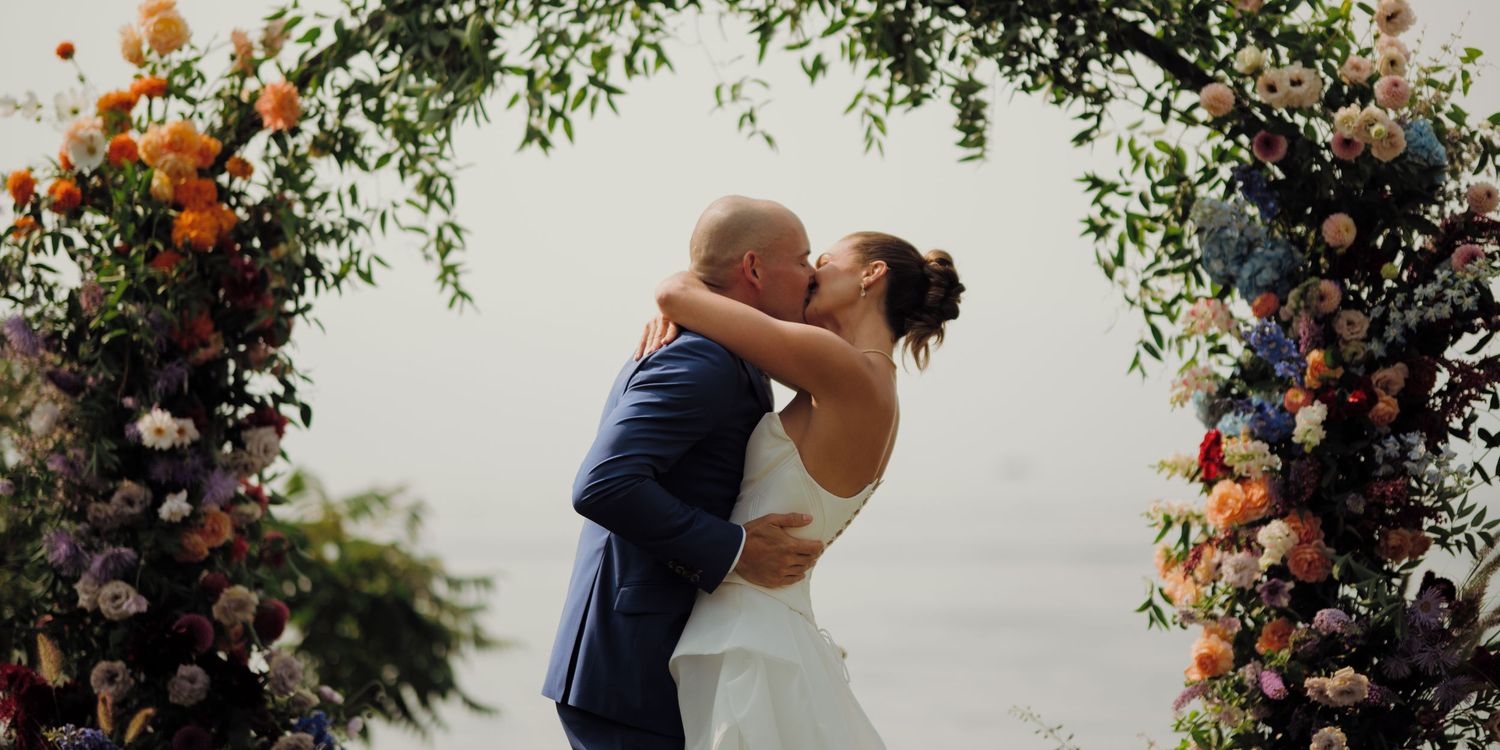 A couple shares a romantic kiss under a beautiful floral arch with orange and pink blooms at an outdoor wedding ceremony.