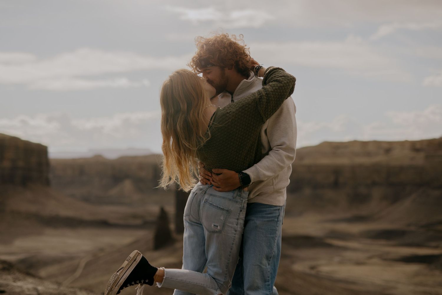 Two people embrace while standing in a scenic Utah desert landscape with dramatic cloudy skies at sunset.