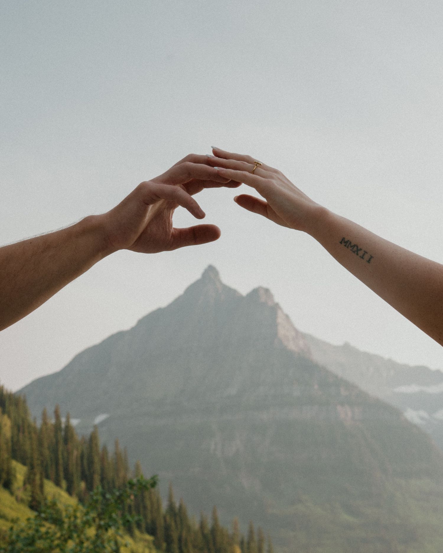 Two hands reaching toward each other against a misty mountain backdrop with green forest below.
