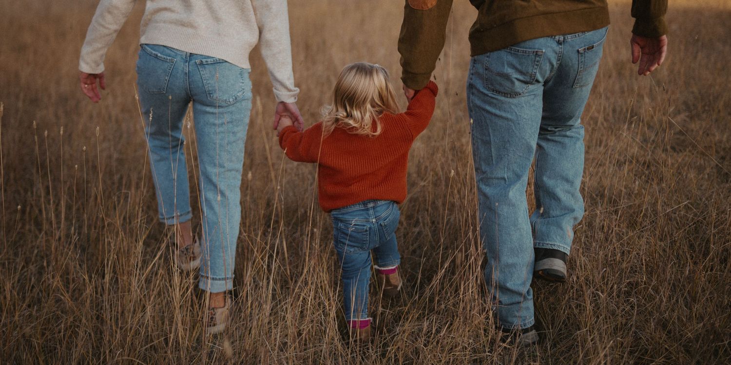 A small child in a red sweater holds hands with two adults while walking through tall brown grass at sunset.