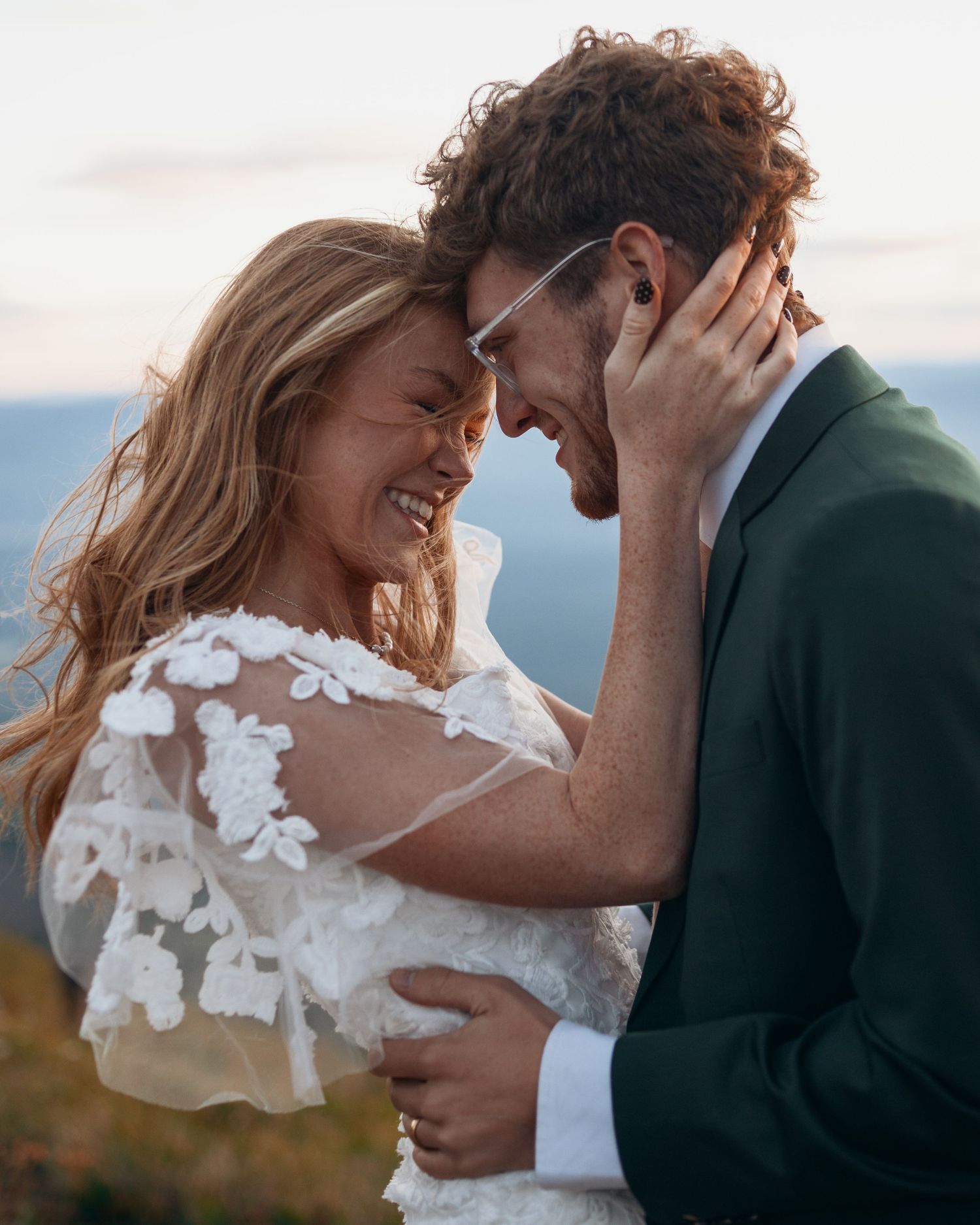 A romantic moment between newlyweds in a white wedding dress and dark suit against a scenic outdoor backdrop.