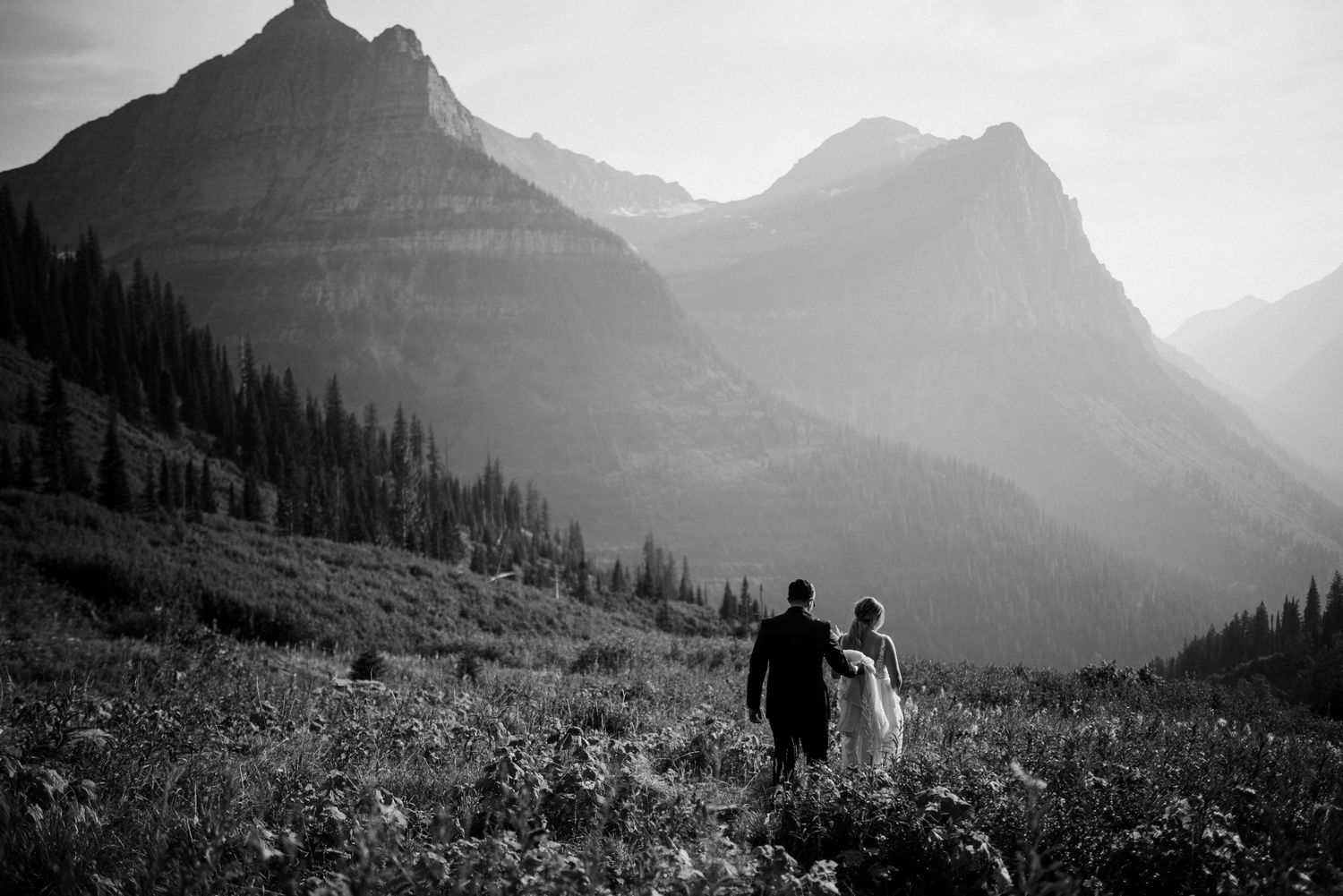 Black and white mountain landscape with silhouettes of hikers walking through a wildflower meadow.