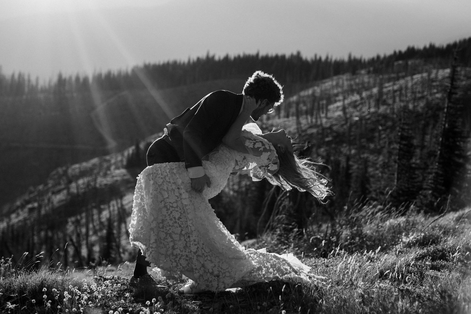 A couple embraces on a mountainside meadow in black and white as sunbeams stream through the misty forest backdrop.