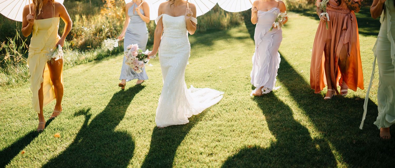 A bride and bridesmaids walking together outdoors in a grassy field during golden hour sunlight.