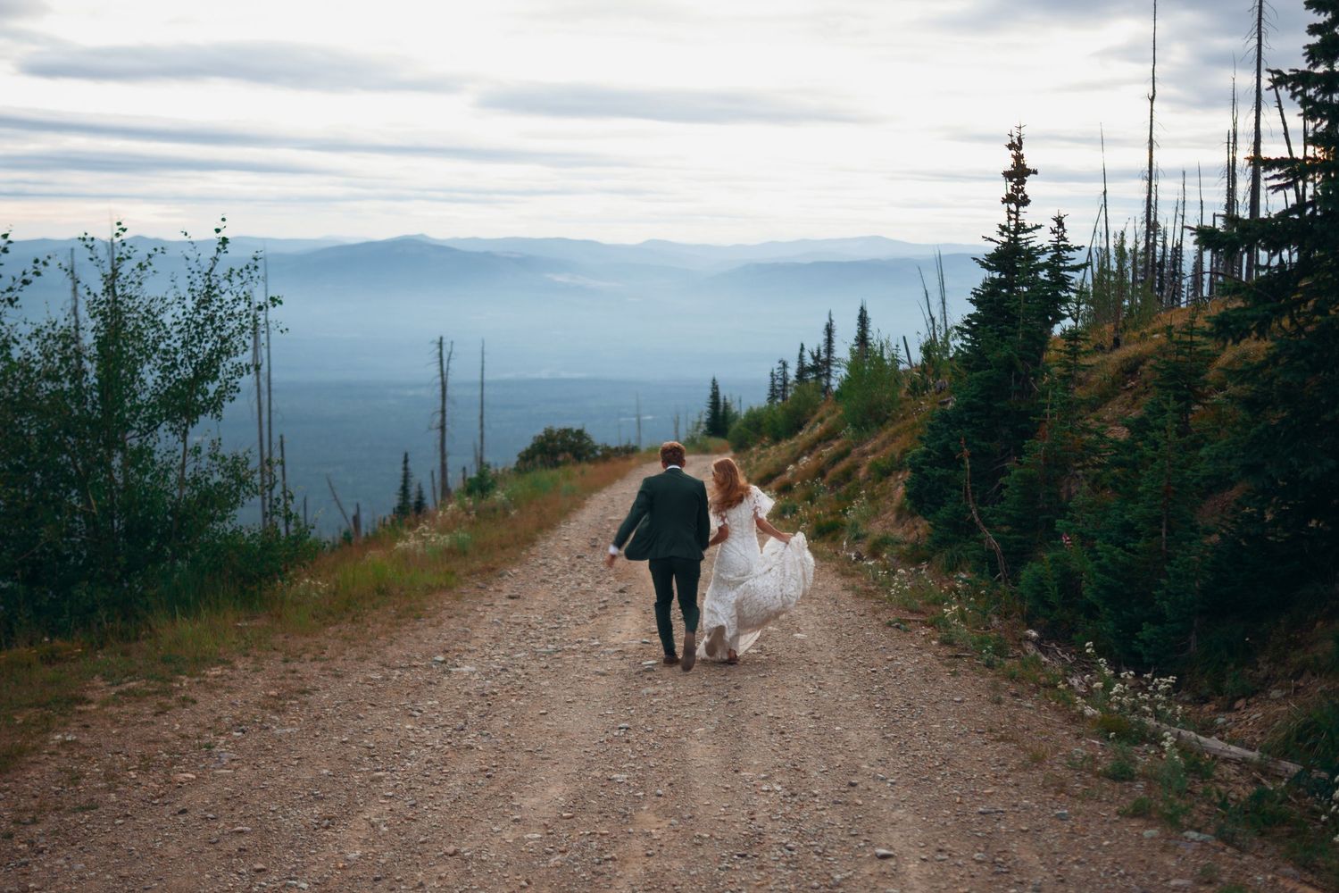 Two people walk along a scenic mountain trail overlooking a vast landscape with evergreen trees and cloudy skies.