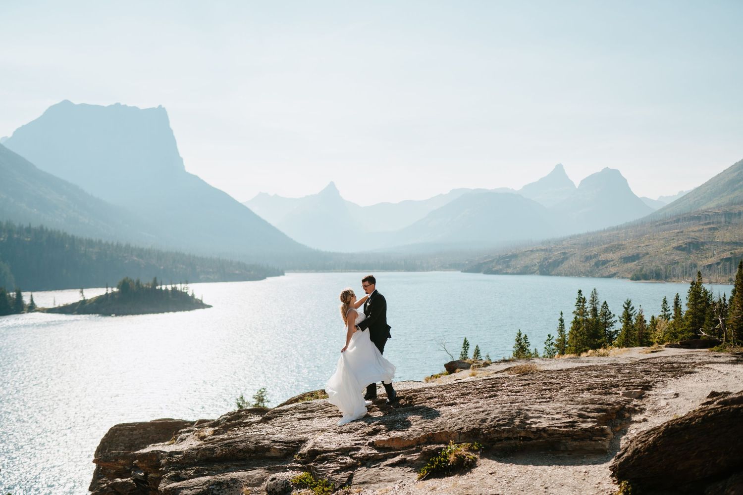 A couple stands on a rocky overlook above a serene mountain lake surrounded by peaks in Glacier National Park.