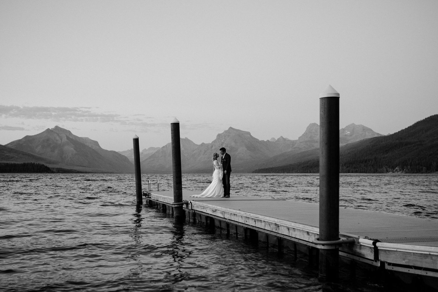 A black and white photograph showing a dock on a mountain lake with a bride and groom silhouetted against Lake McDonald's mountain scape.