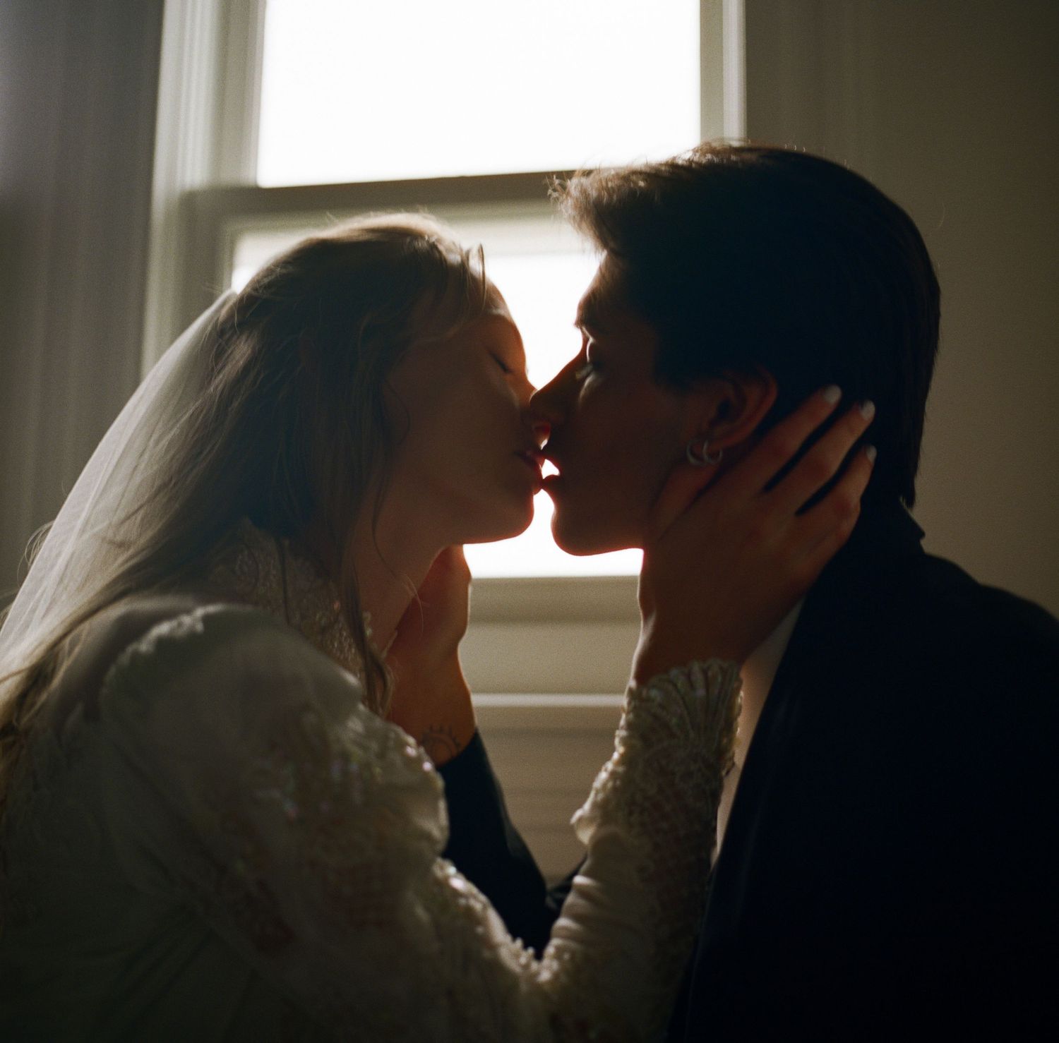 Bride and groom sharing a tender kiss near a sunlit window on their wedding day.