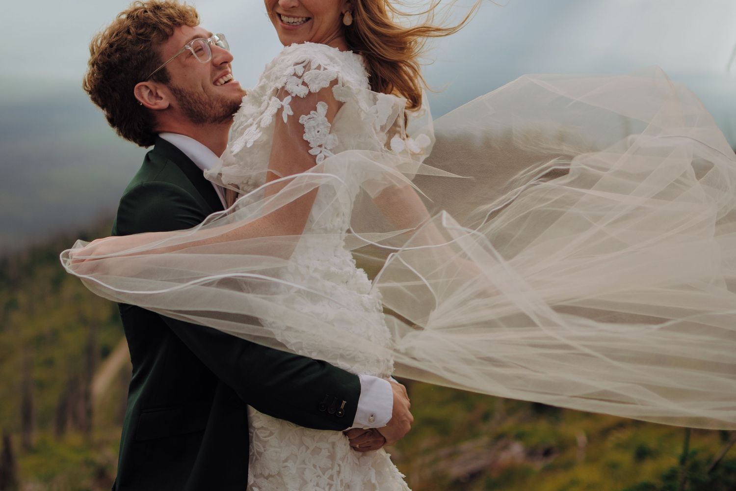 A romantic wedding couple embraces outdoors while the bride's veil flows dramatically in the wind.