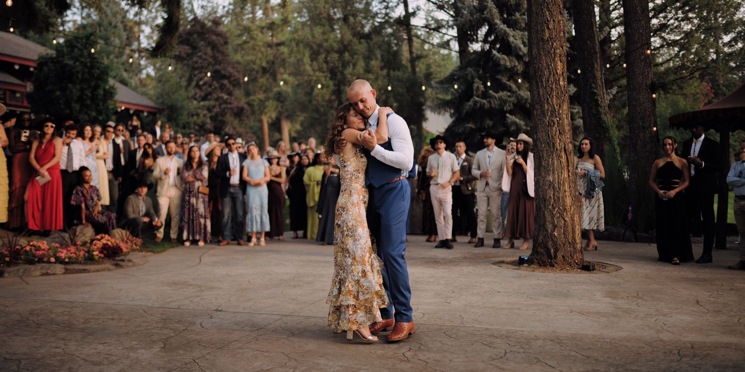 A couple shares their first dance at an outdoor wedding reception as guests watch from beneath tall pine trees at dusk.