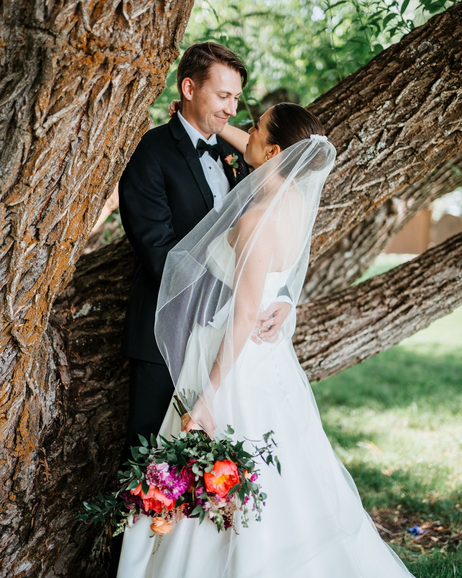 Wedding couple poses romantically under large tree branches with colorful floral bouquet.