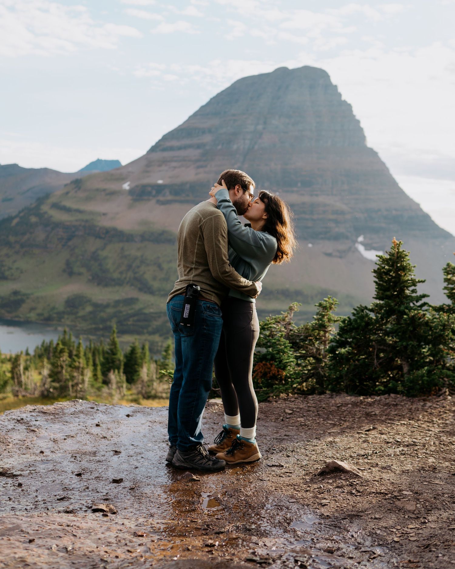 Couple sharing a romantic kiss on a mountain overlook with a dramatic peak and alpine lake in the background.