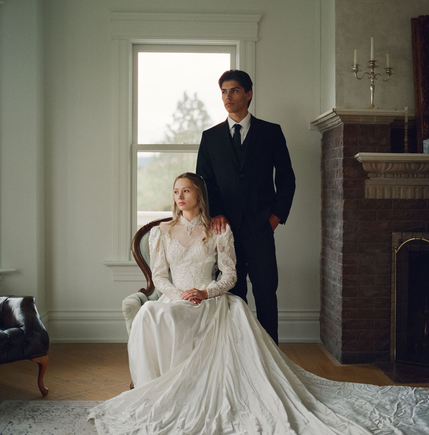 Bride in white lace gown seated in chair with groom in dark suit standing beside her near fireplace.