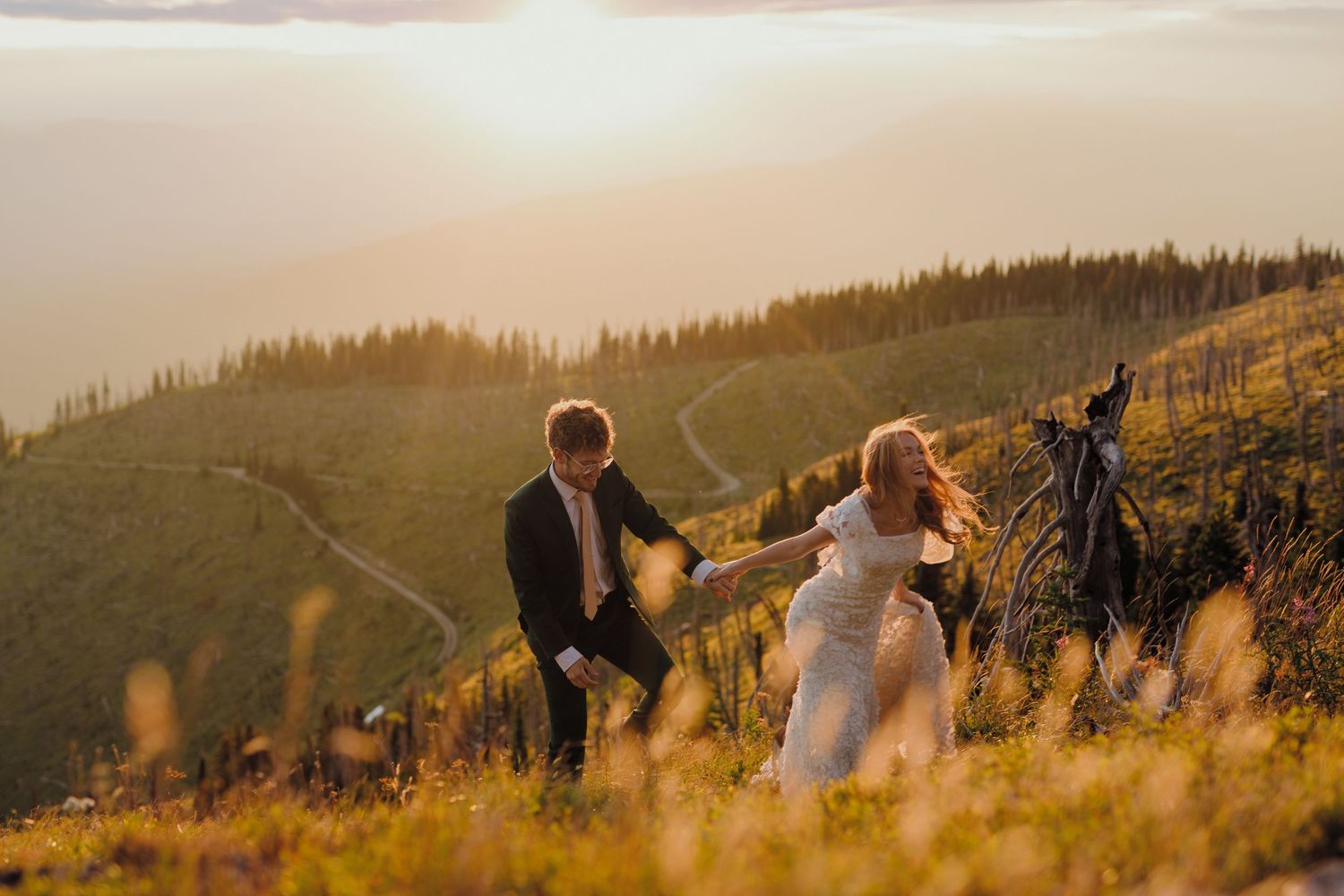 Two people stand together on a golden hillside at sunset with mountains and forest in the background.