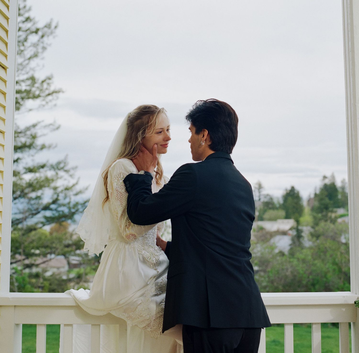 A bride and groom share a romantic moment on a white porch, with the bride in a veil and groom in a black suit.