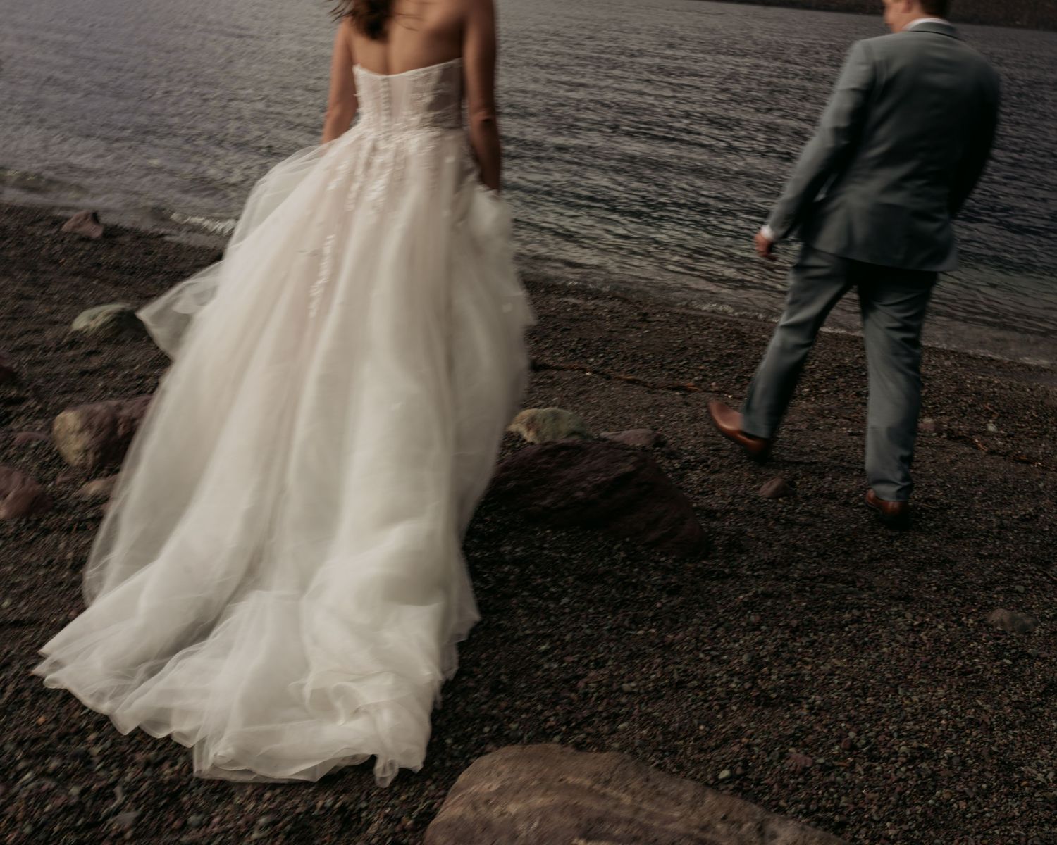 A bride in a large, flowing white wedding dress stands on a rocky, rural landscape with a man in a suit nearby.